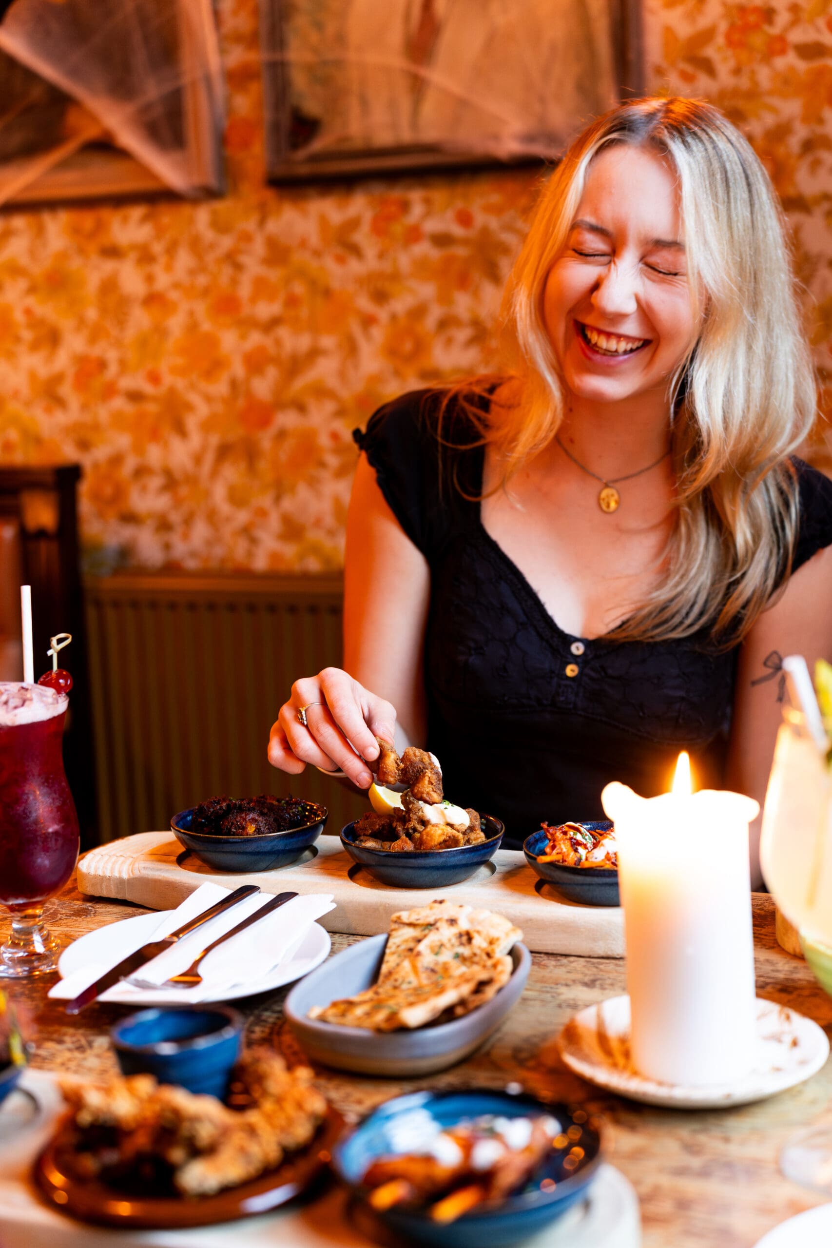 A smiling woman with long blonde hair sits at a table in a cozy lounge, reaching for food. A lit candle and colorful drinks add warmth and charm to the inviting dining atmosphere.