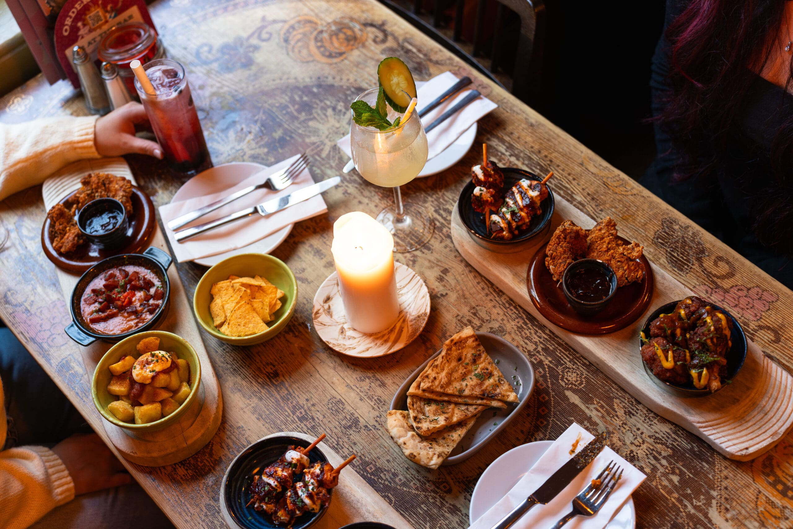 A rustic wooden table set with plates of tapas, nachos, bread, fried appetizers, dips, drinks, and a lit candle at the center creates a cozy lounge vibe, surrounded by people ready to eat.