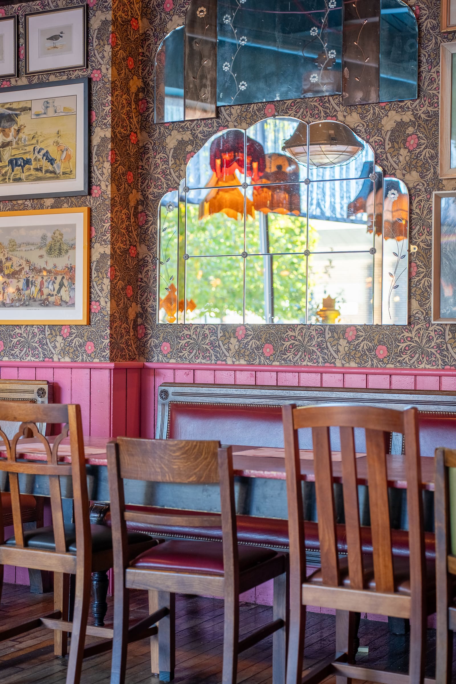 A vintage-style dining area with wooden chairs and a bench, floral wallpaper, framed artwork, and a decorative mirror reflecting orange lamps and greenery outside—perfect for displaying a Tarro centerpiece.