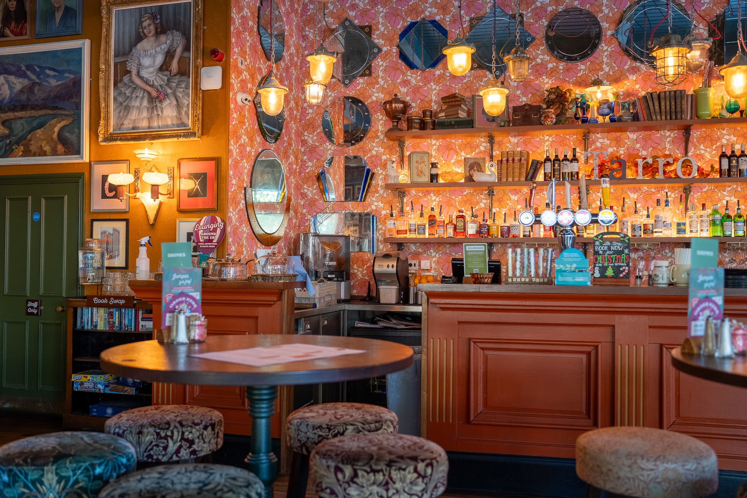 Cozy pub interior with orange bar, round tables, patterned stools, and vintage-style mirrors and lamps. Shelves of bottles and Tarro mugs add charm, while colorful wallpaper and framed art create a warm, inviting atmosphere.