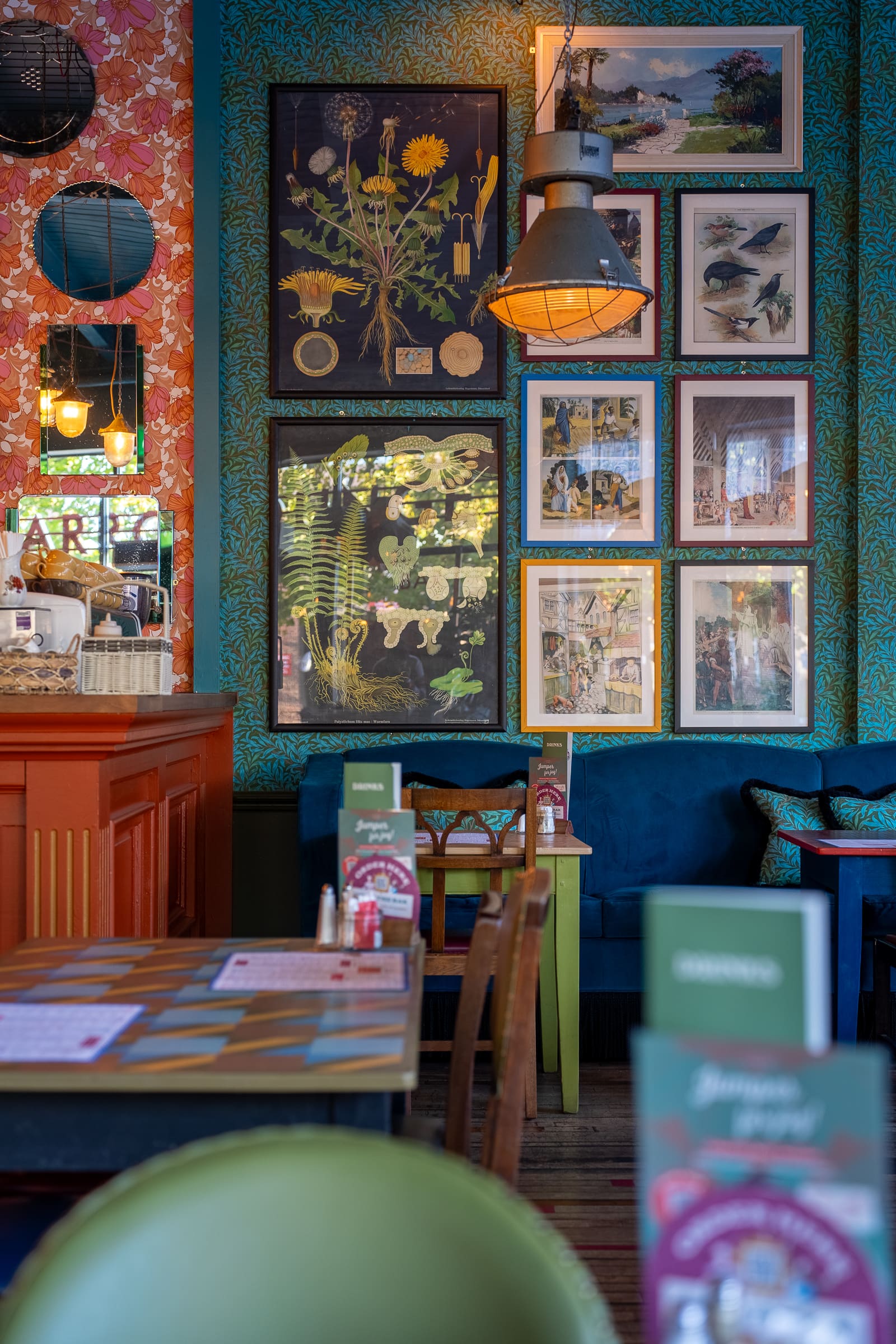 A cozy café interior with colorful walls, Tarro jars on tables, botanical and bird-themed framed art, retro pendant lights, a small orange counter, and a blue padded bench lining the far wall.