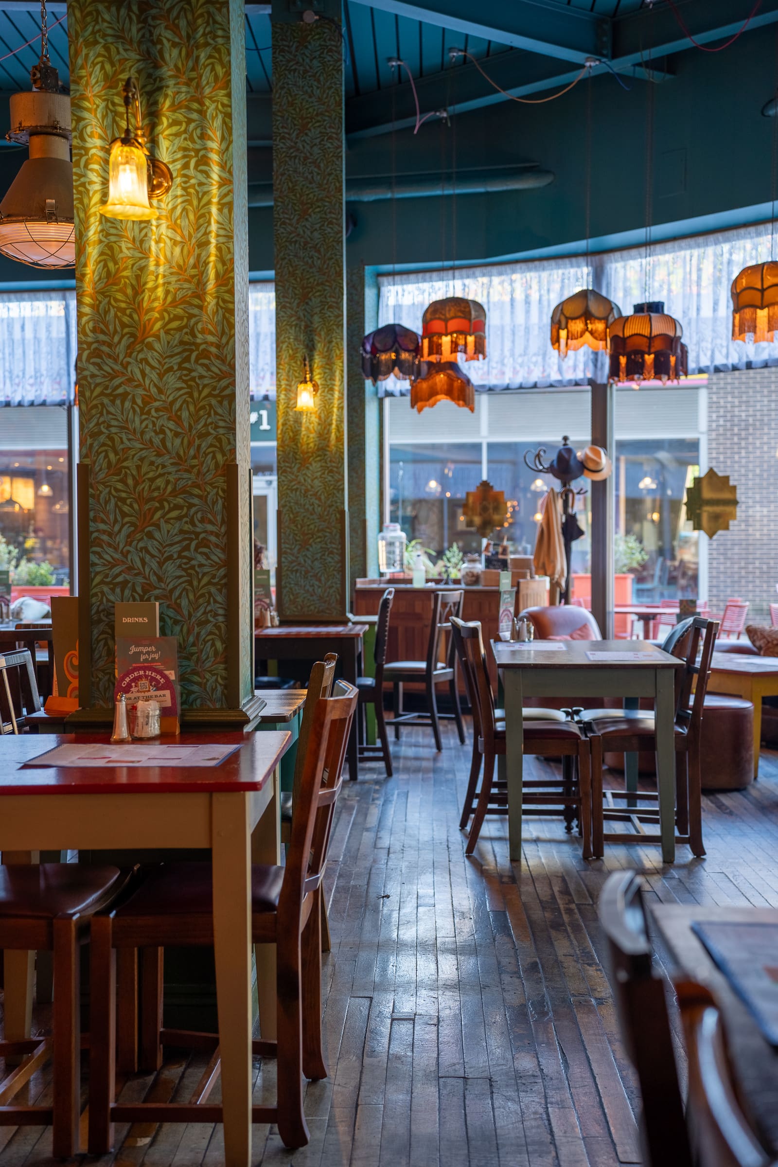 Cozy café interior with colorful vintage lamps, patterned green wallpaper, and wooden tables and chairs. Large windows let in natural light, with some tables set for customers—each adorned with a charming Tarro. The atmosphere is warm and inviting.