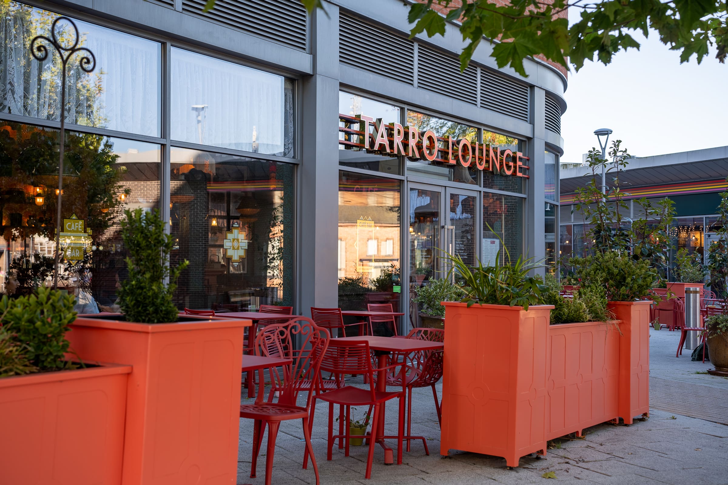 Outdoor seating area with red chairs and tables in front of Tarro Lounge. Large orange planters with greenery line the patio, complementing the café’s modern exterior and spacious windows.
