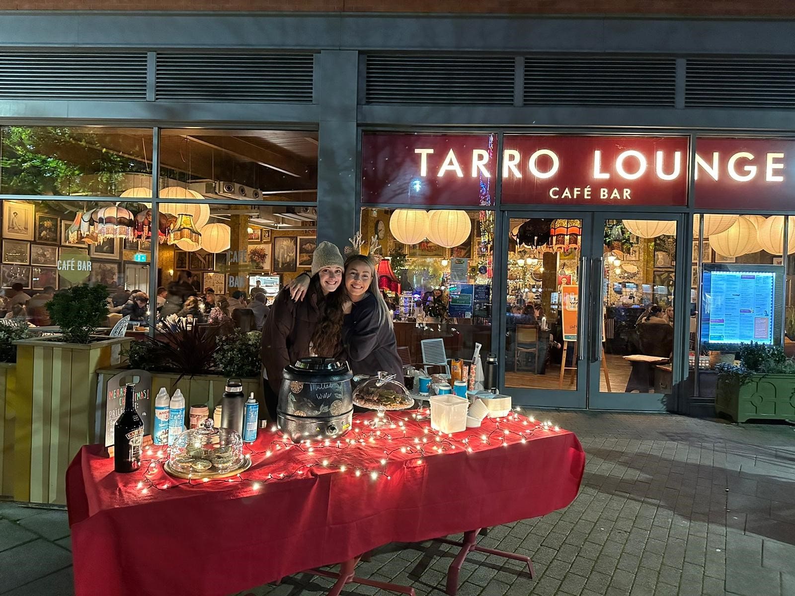 Two people smile behind a red table covered with treats, drinks, and fairy lights, set up outside Tarro Lounge Café Bar at night, with people dining inside the brightly lit café.
