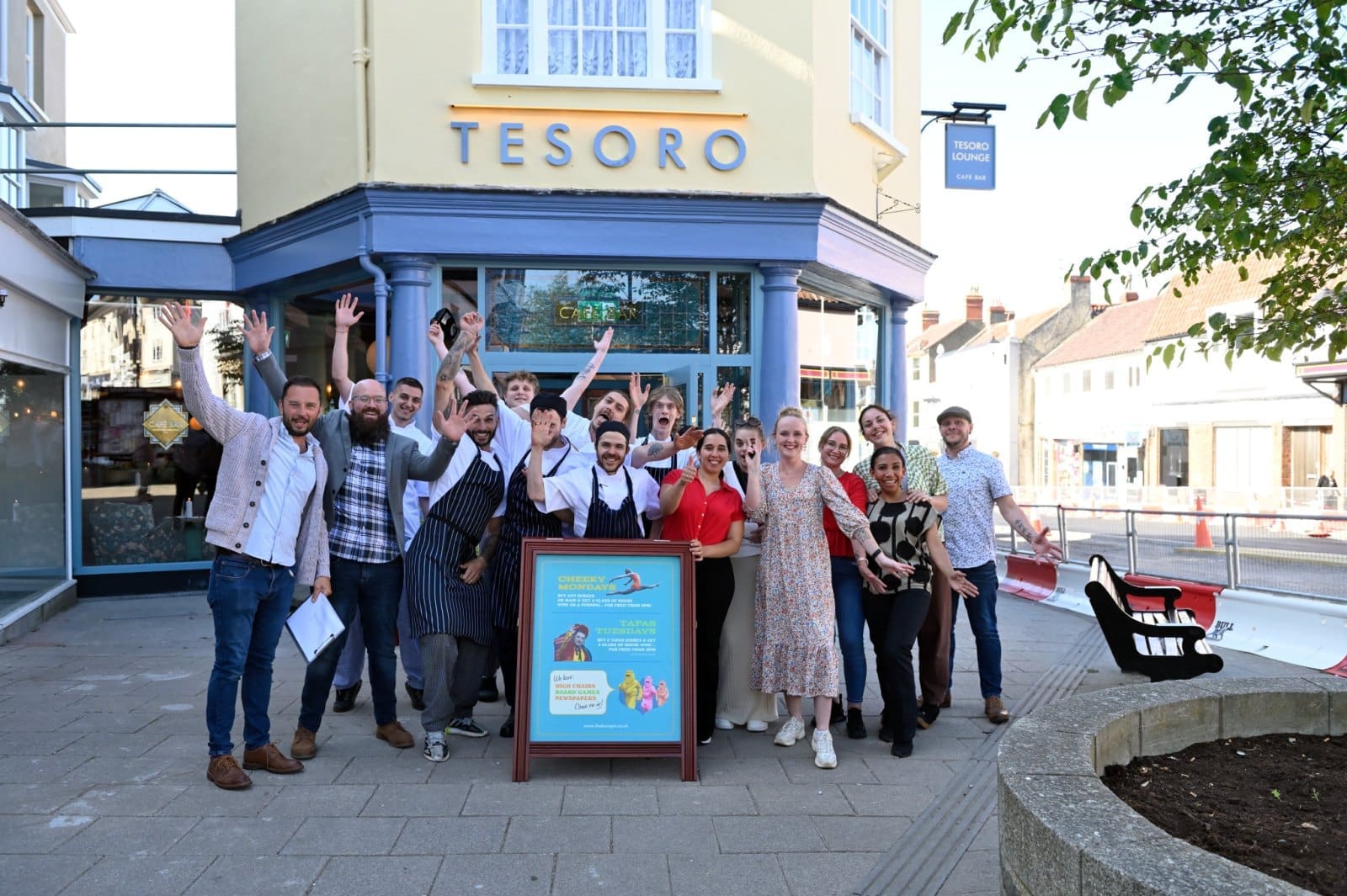 A group of people, including restaurant staff in Bianco uniforms, stand smiling and waving outside a building with a sign that reads "TESORO." A colorful signboard is displayed in front of the group on the sidewalk.