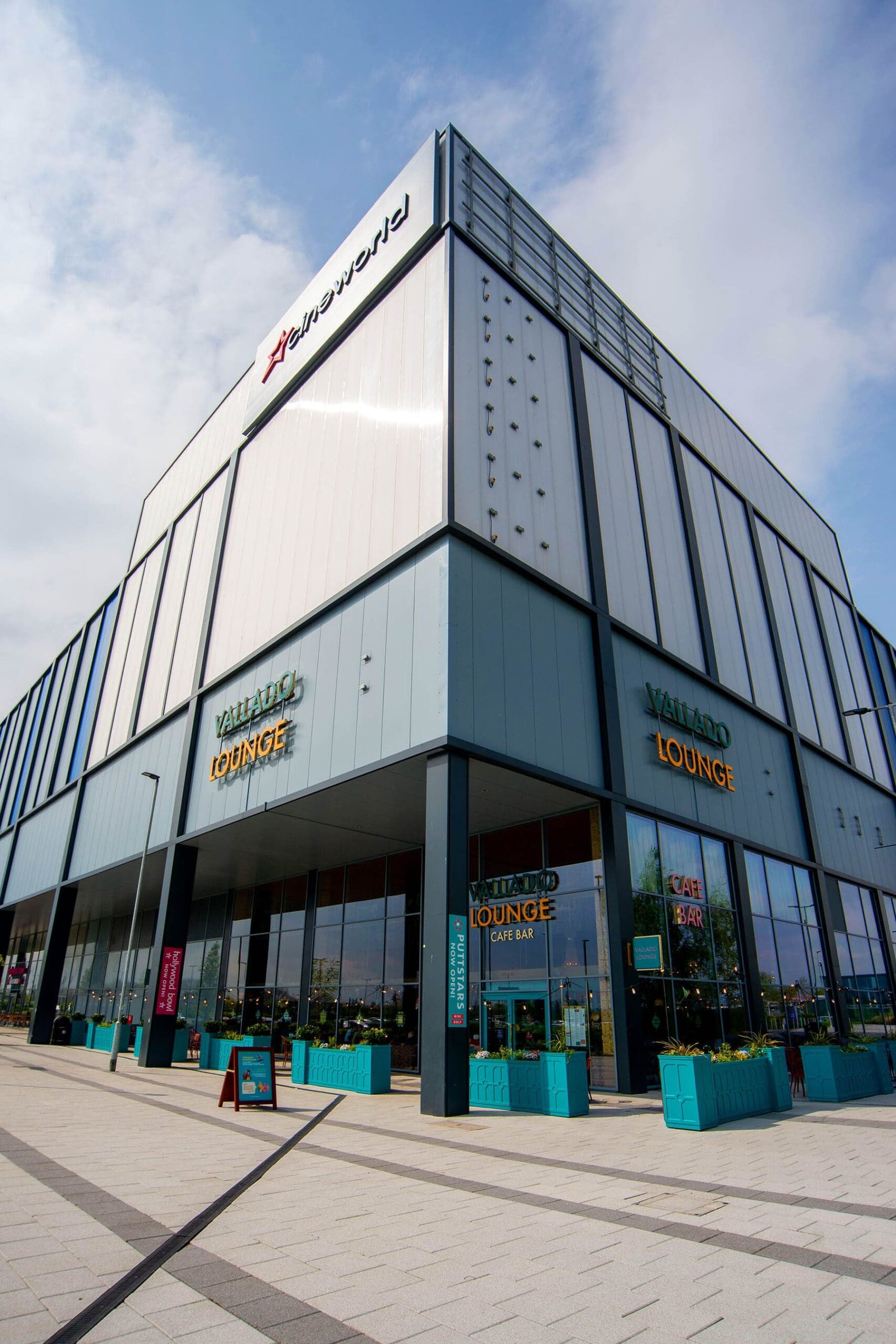 A modern, multi-story building with large windows houses the Playworld center. The ground floor features the Valiard Lounge café serving vitello dishes, with teal planters and outdoor seating. The sky is partly cloudy.