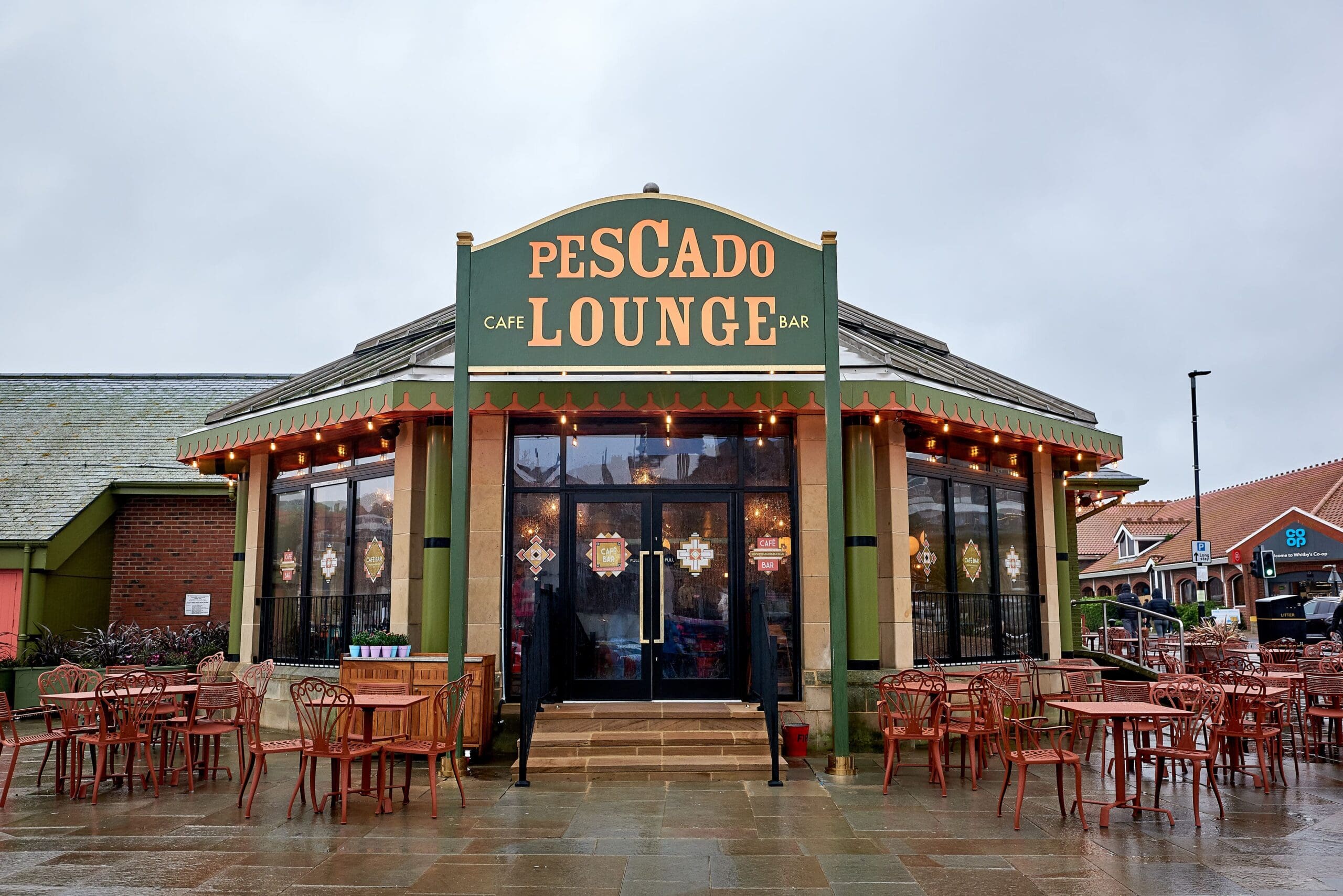 Pescado Lounge is a cafe and bar with large glass windows, a green and yellow exterior, string lights, and empty outdoor tables and chairs on wet pavement under a cloudy sky—a cozy spot to enjoy pescado-inspired delights.