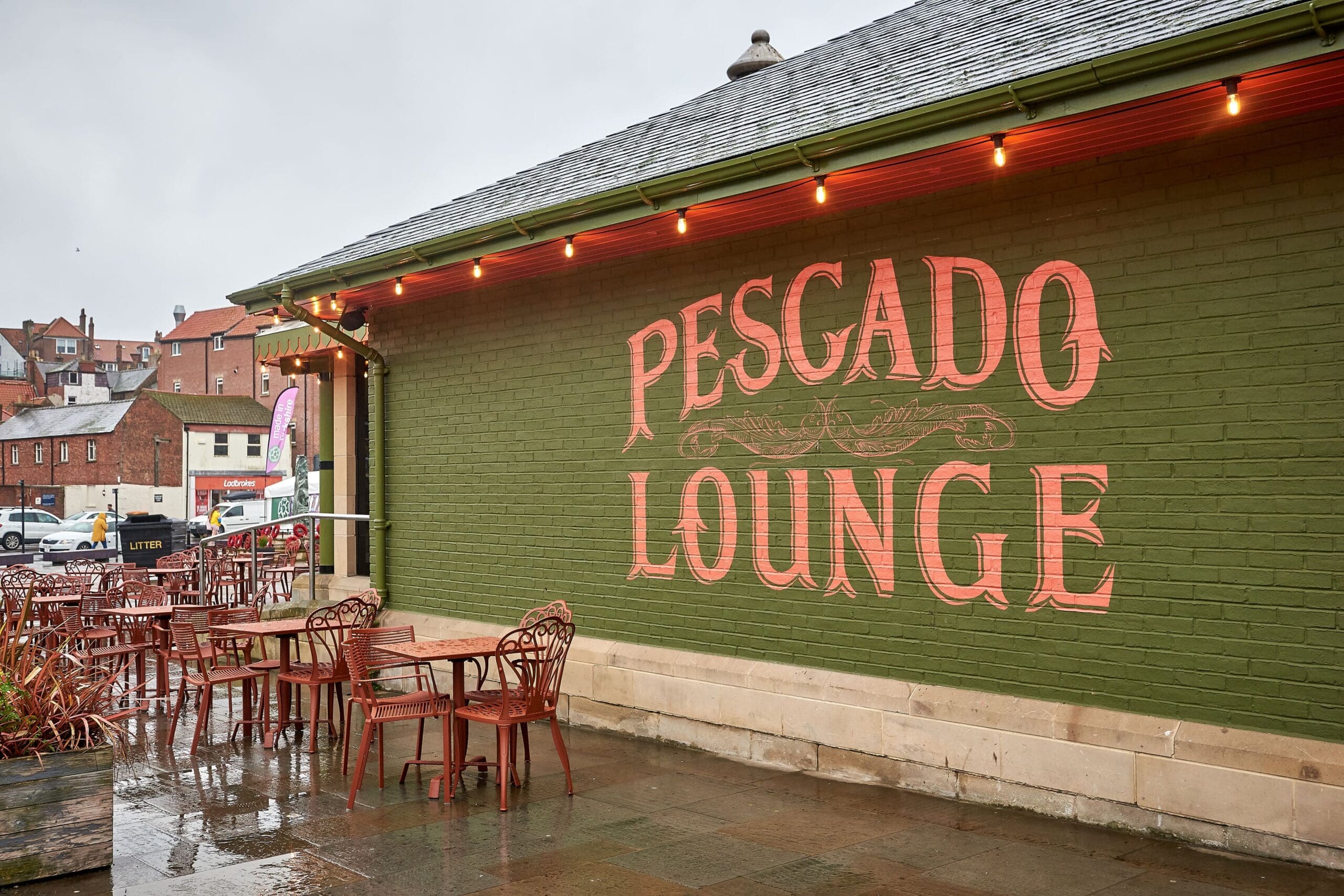 Outdoor café with empty red chairs and tables on a wet pavement beside a green brick building. Large painted letters on the wall read "Pescado Lounge," adding character to the scene under an overcast sky with buildings in the background.