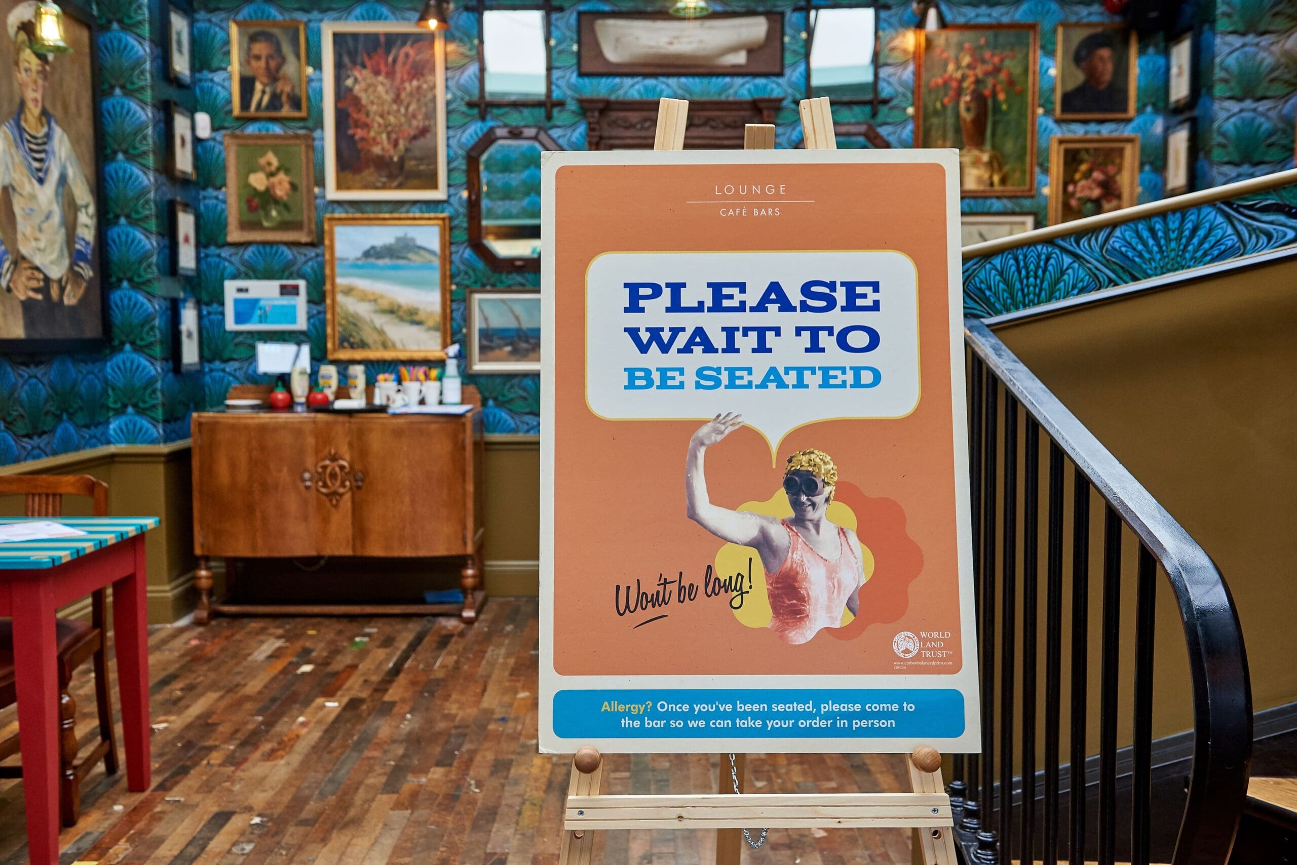 A sign in a stylish, art-filled café reads, "PLEASE WAIT TO BE SEATED," featuring a vintage-style pescado illustration and colorful decor in the background, including framed paintings and patterned wallpaper.