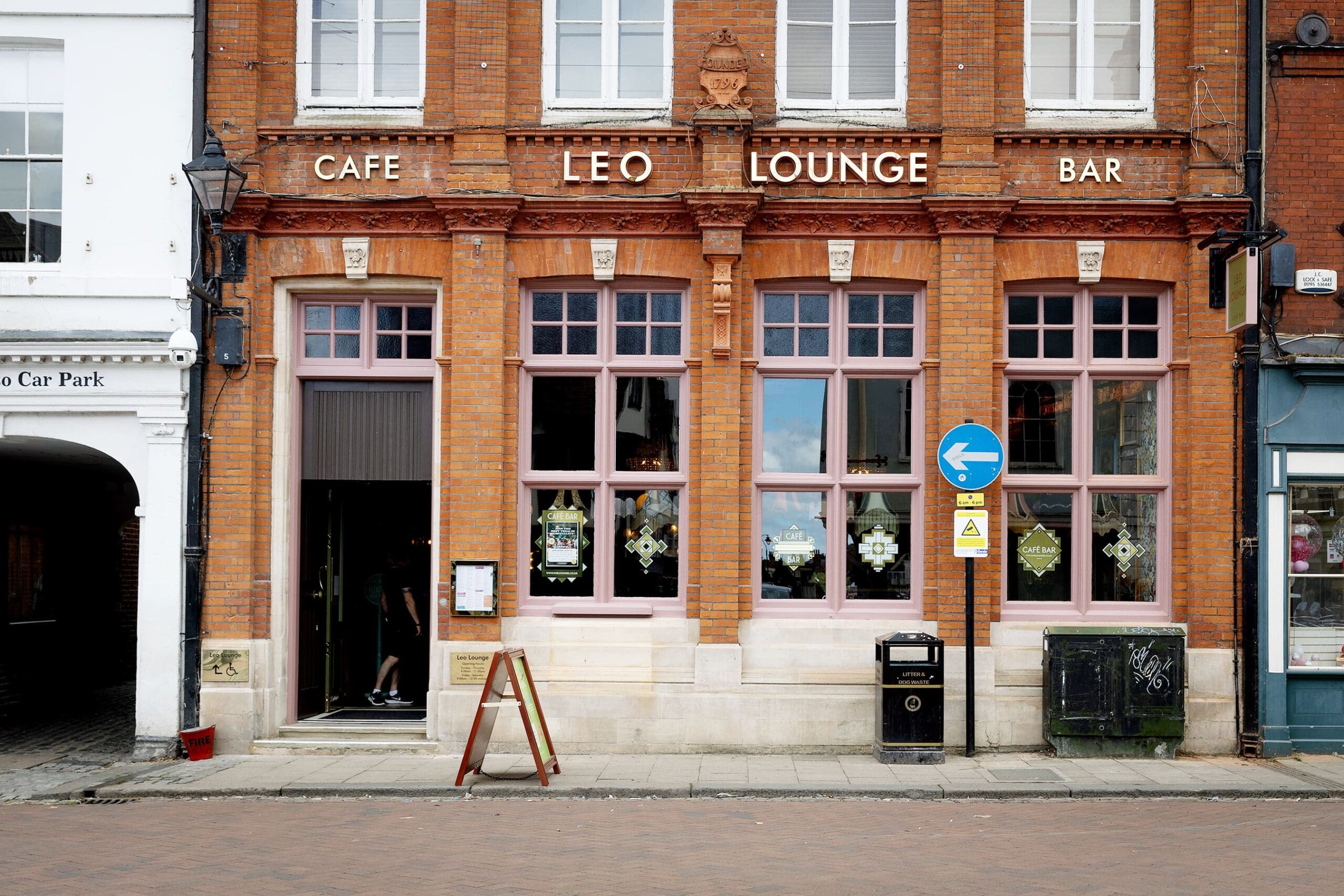 Street view of a brick building with large windows and signs reading "CAFE LEO LOUNGE BAR." Leo’s open door, menu board, trash bin, and various street signs are visible in front.