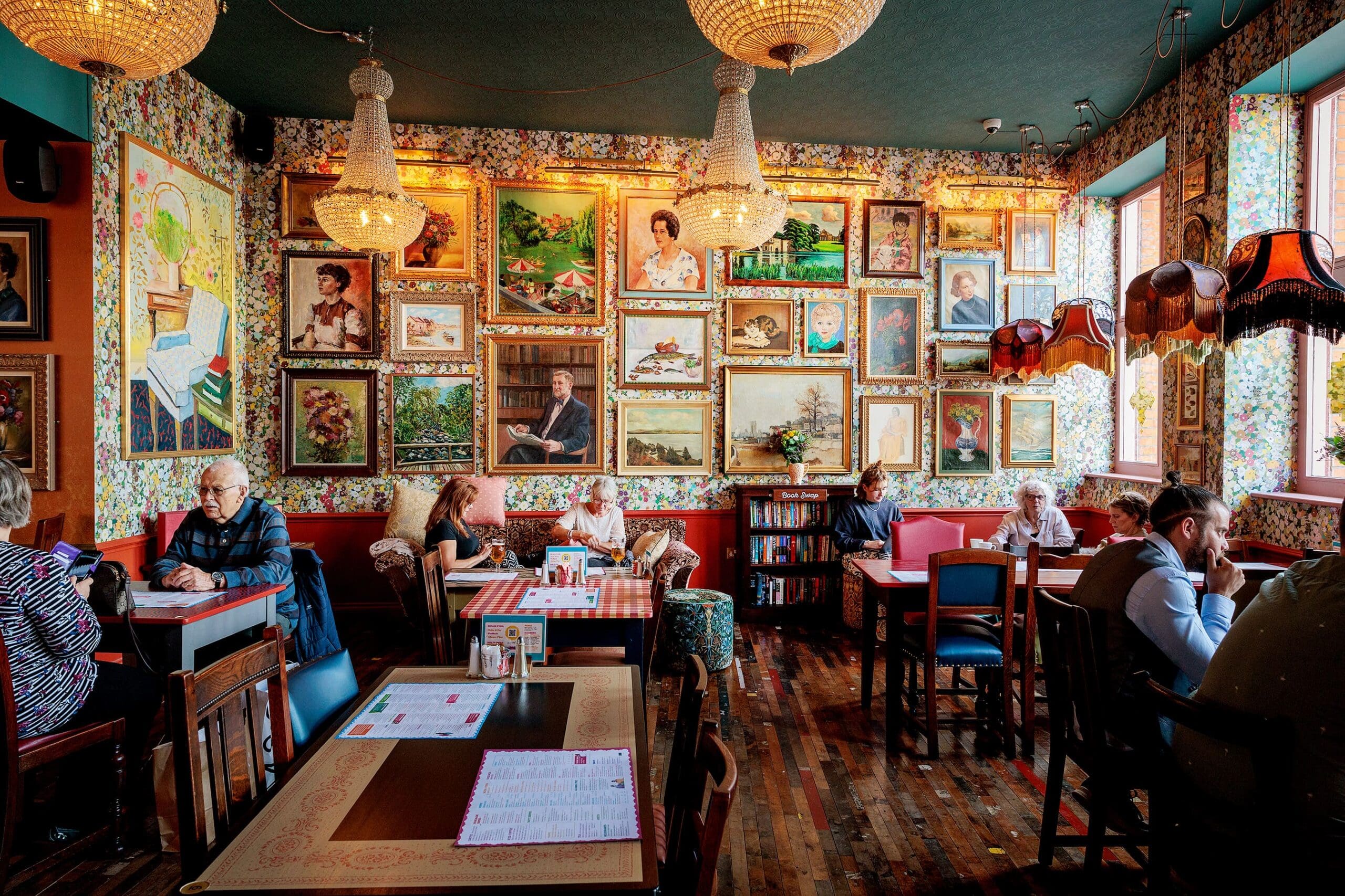 A cozy, eclectic restaurant with patterned wallpaper and many framed paintings—some featuring majestic leos—adorn the walls. Several people sit at tables, chatting or reading menus, while warm light from chandeliers fills the room.