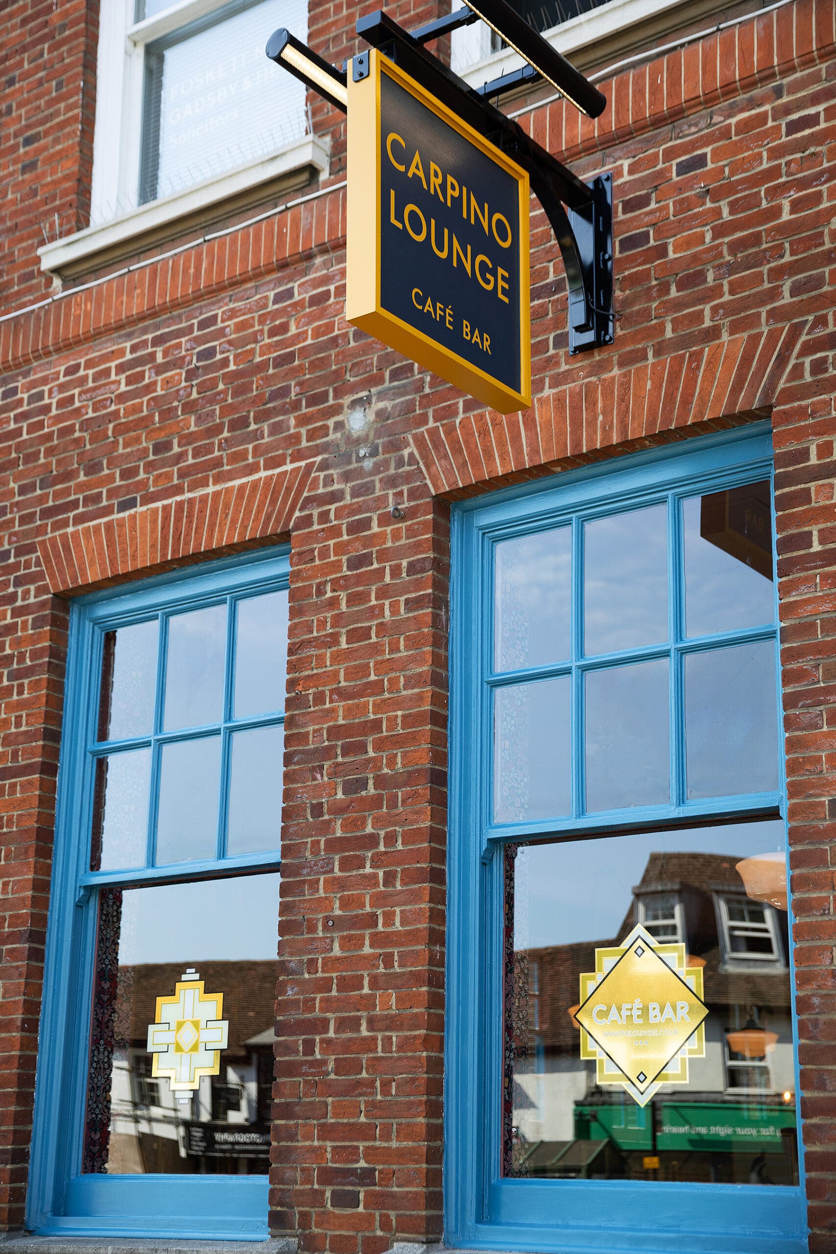 A brick building with two blue-trimmed windows and a hanging yellow-and-black sign reading "Carpino Lounge Café Bar." The windows display geometric yellow carpino café bar decals.
