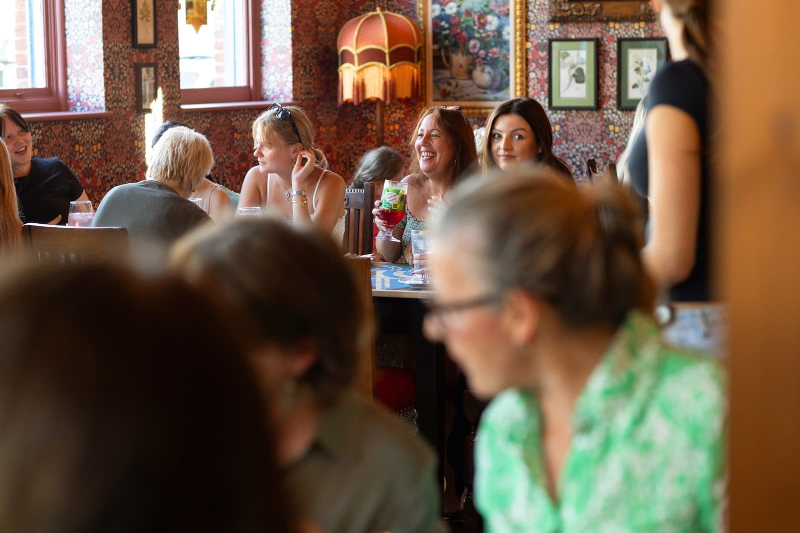 A group of women sit and talk at a lively, warmly lit café or restaurant—perhaps Carpino—with colorful wallpaper and framed art in the background. Some smile and hold drinks, while one woman in green is blurred in the foreground.