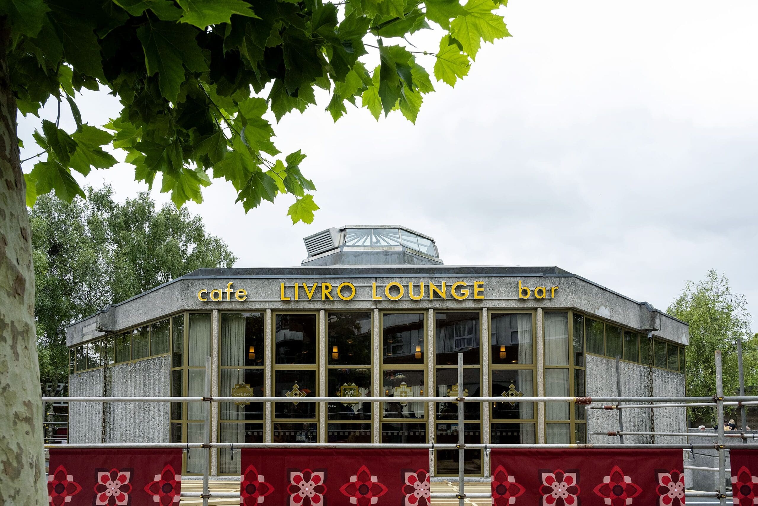 A modern café named Livro Lounge features large glass windows and gold accents, with its sign "LIVRO LOUNGE" above the entrance. Green trees and red decorative barriers add charm to the inviting scene.