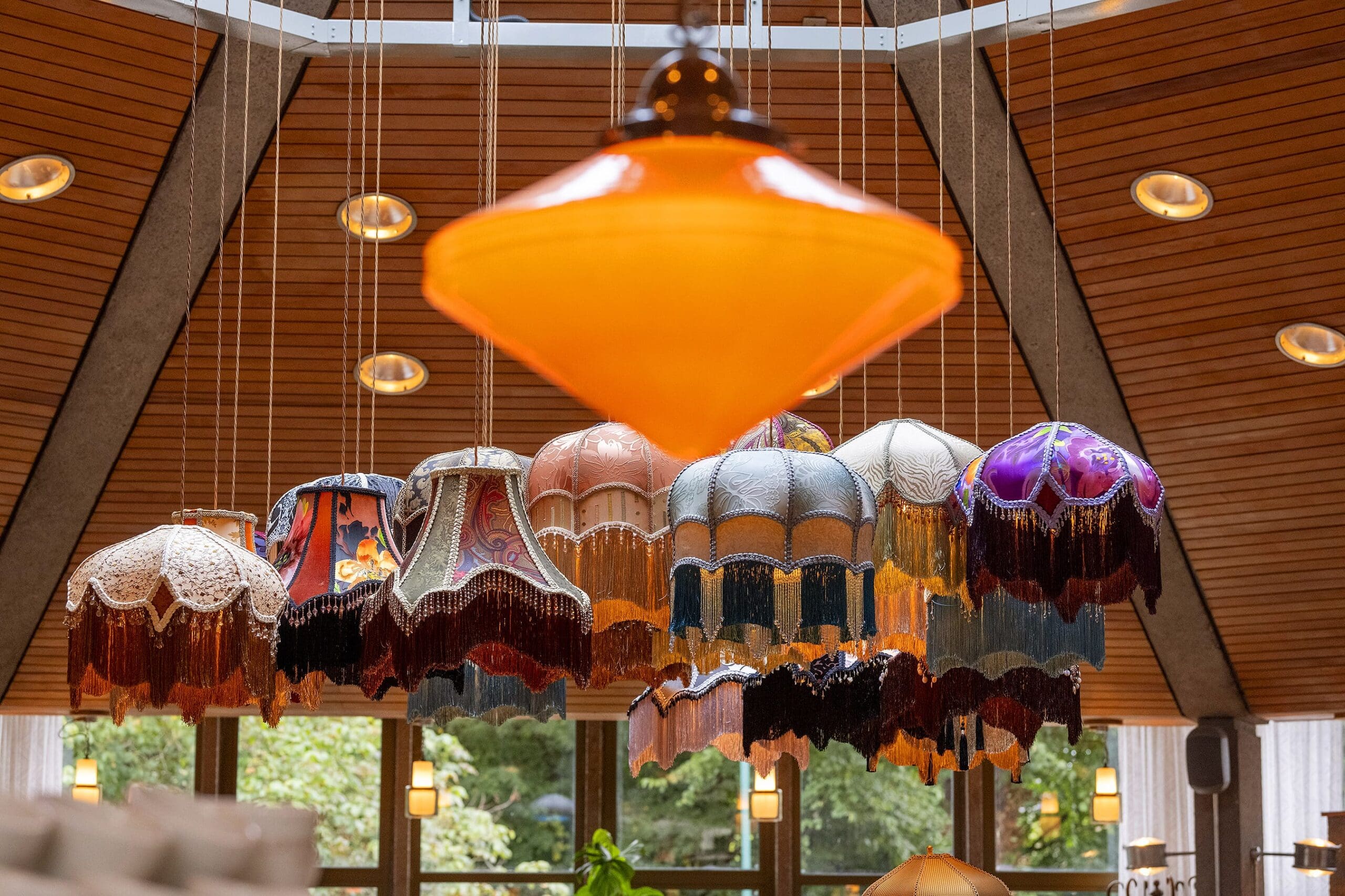 A group of colorful, vintage-style lampshades with fringe hang from the ceiling beneath wooden panels, while a Livro rests nearby and windows and greenery are visible in the background.