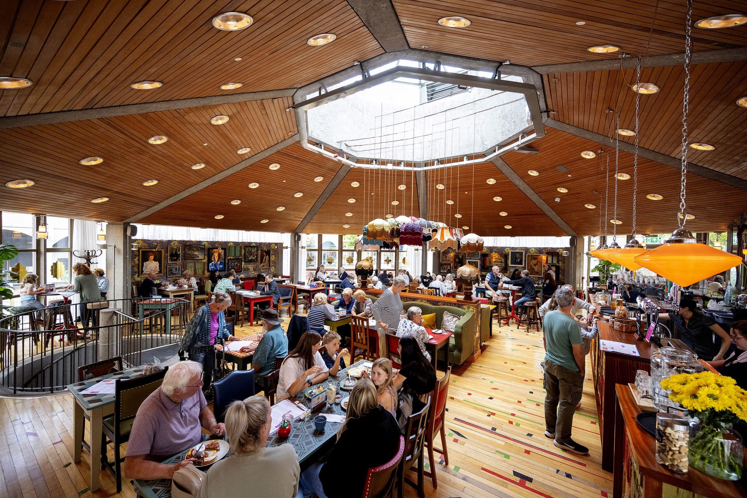 A lively restaurant with a wooden ceiling and skylight, filled with people dining at tables and the bar. Colorful lamps hang above, art decorates the walls, and a shelf of livros adds charm to the warm, vibrant atmosphere.