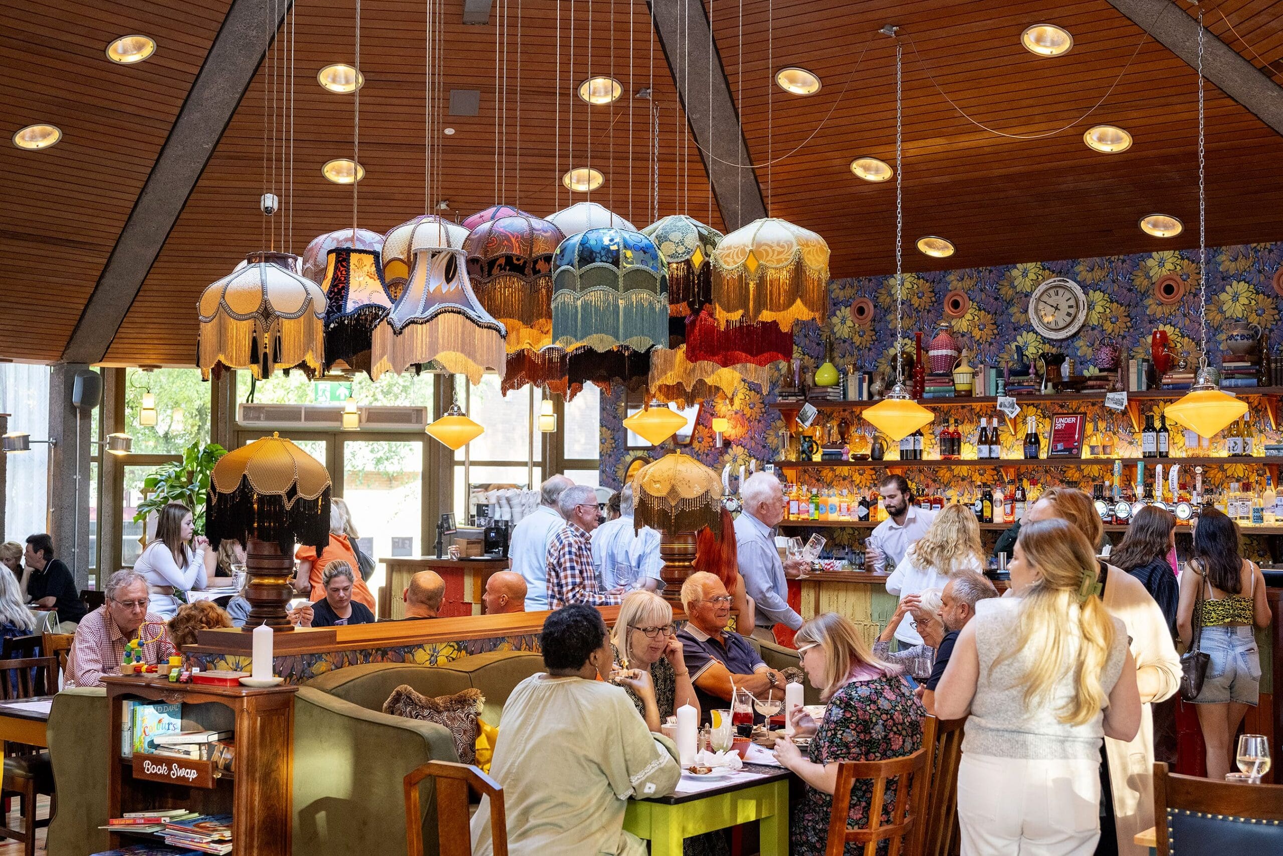 A lively restaurant with vintage lamps, floral wallpaper, and people dining and socializing—some flipping through a Livro at their tables. Warm lighting and colorful decor create a cozy, bustling atmosphere.