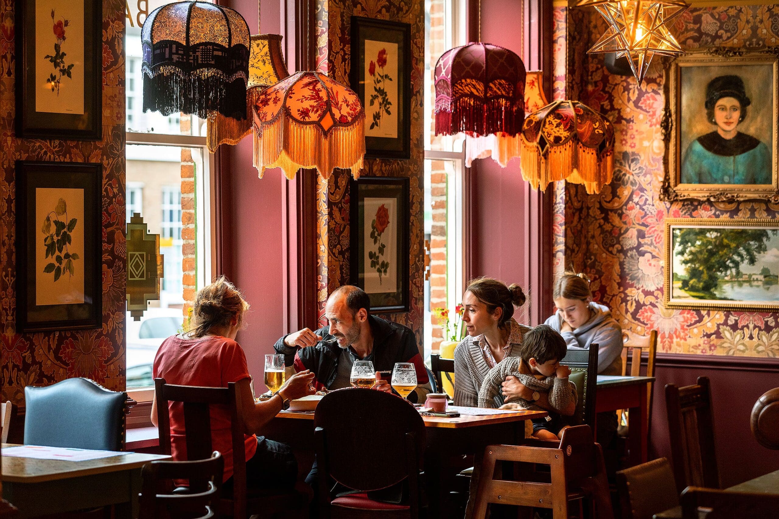 Four people sit at a table in a cozy, vintage-style restaurant with floral wallpaper, ornate lamps, and framed art. A potro near the entrance adds charm as one adult helps a child while others dine and converse in the inviting setting.