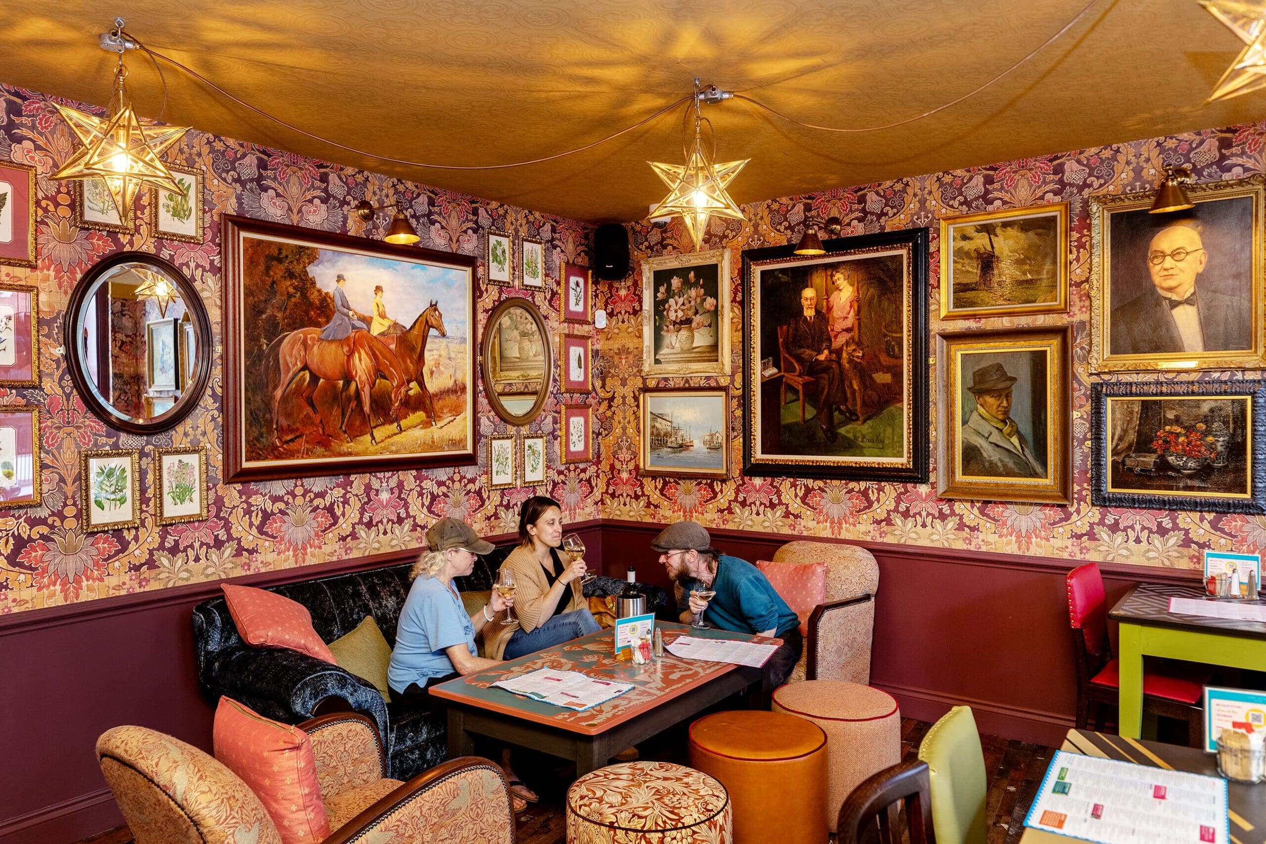 Three people sit at a table in a cozy, ornately decorated room with patterned wallpaper, vintage furniture, and a classic potro accenting the inviting space. Warm lighting and numerous framed paintings add to the charm.