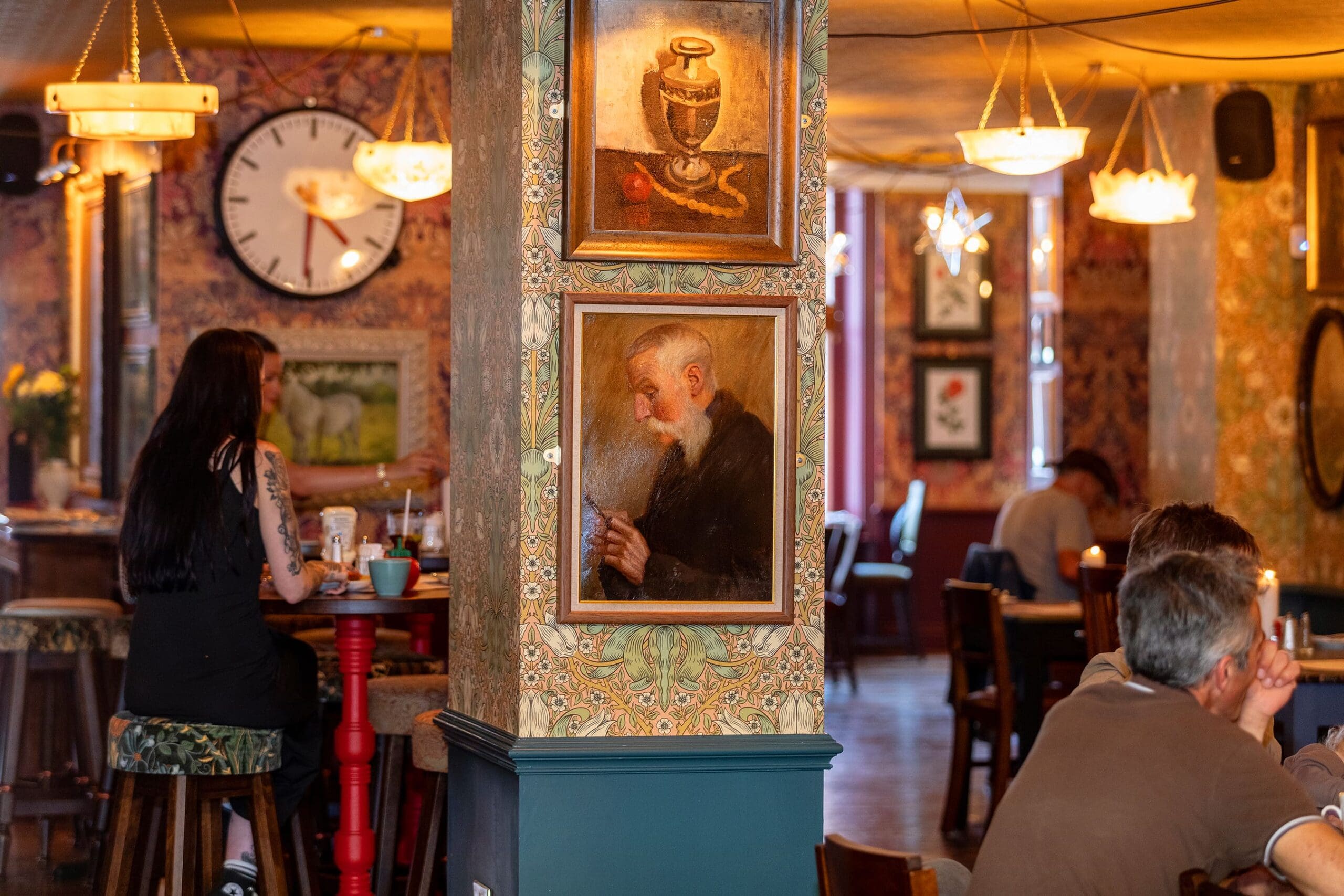 A cozy cafe interior with warm lighting, people sitting at tables, and framed artwork on the walls. A large clock and paintings, including one of a potro, are visible on a column in the center of the image.