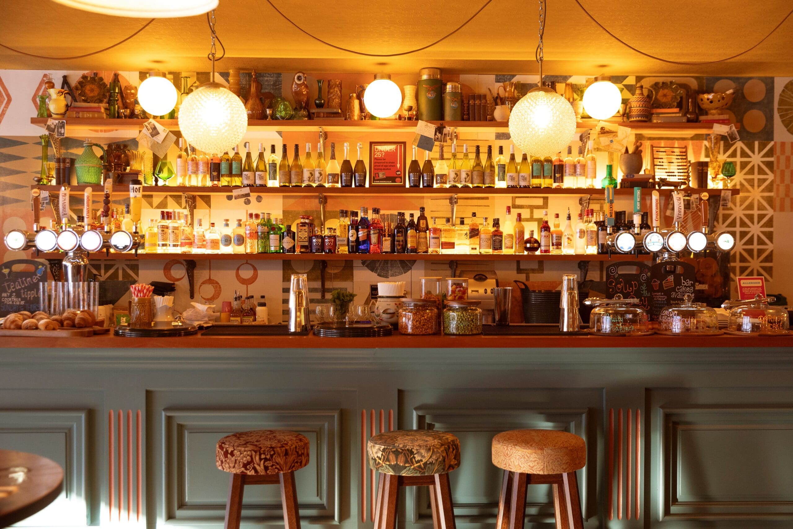 A warmly lit Maggio bar with three stools, shelves lined with bottles, glasses, and decorations, illuminated by hanging globe lights, and a counter displaying pastries and snacks.