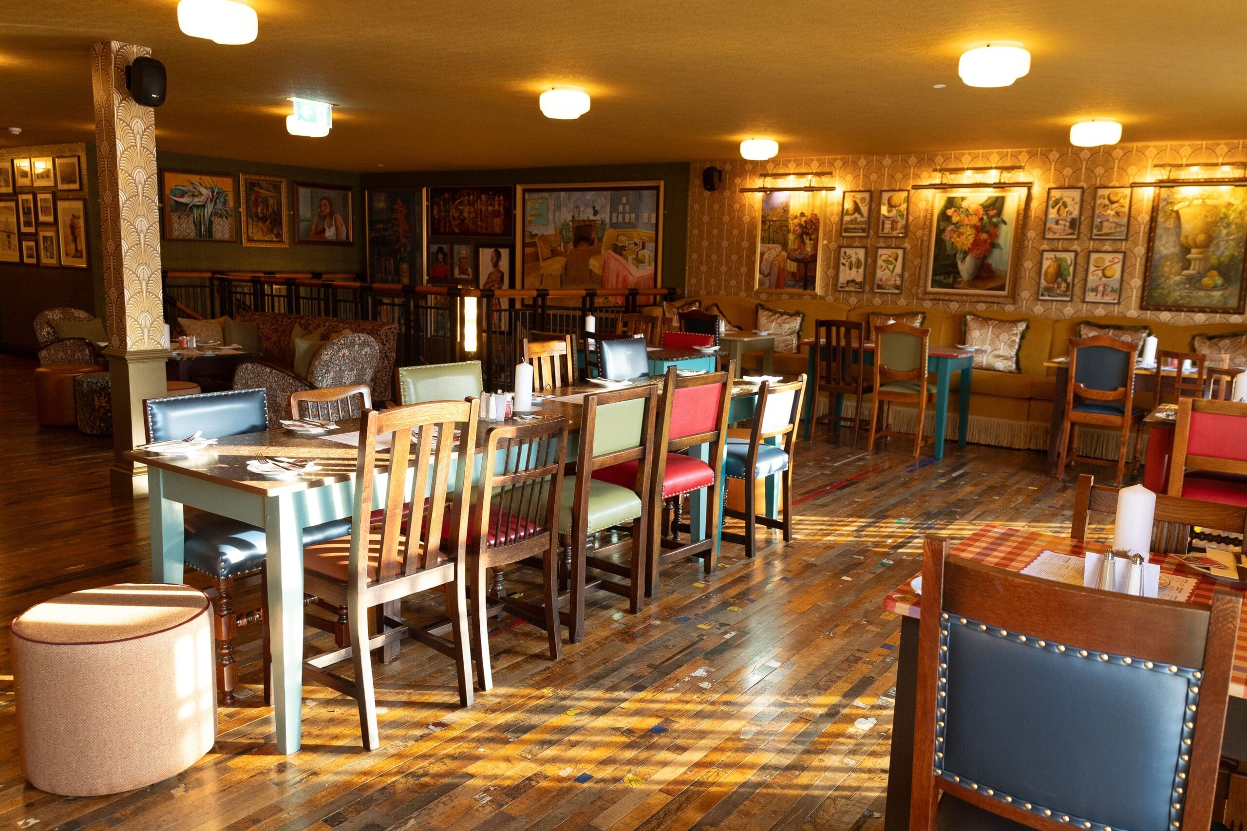 A brightly lit restaurant with wooden floors, colorful chairs, and tables set for dining. Artworks and framed pictures line the warm-toned walls, creating a cozy Maggio atmosphere as sunlight streams in through unseen windows.