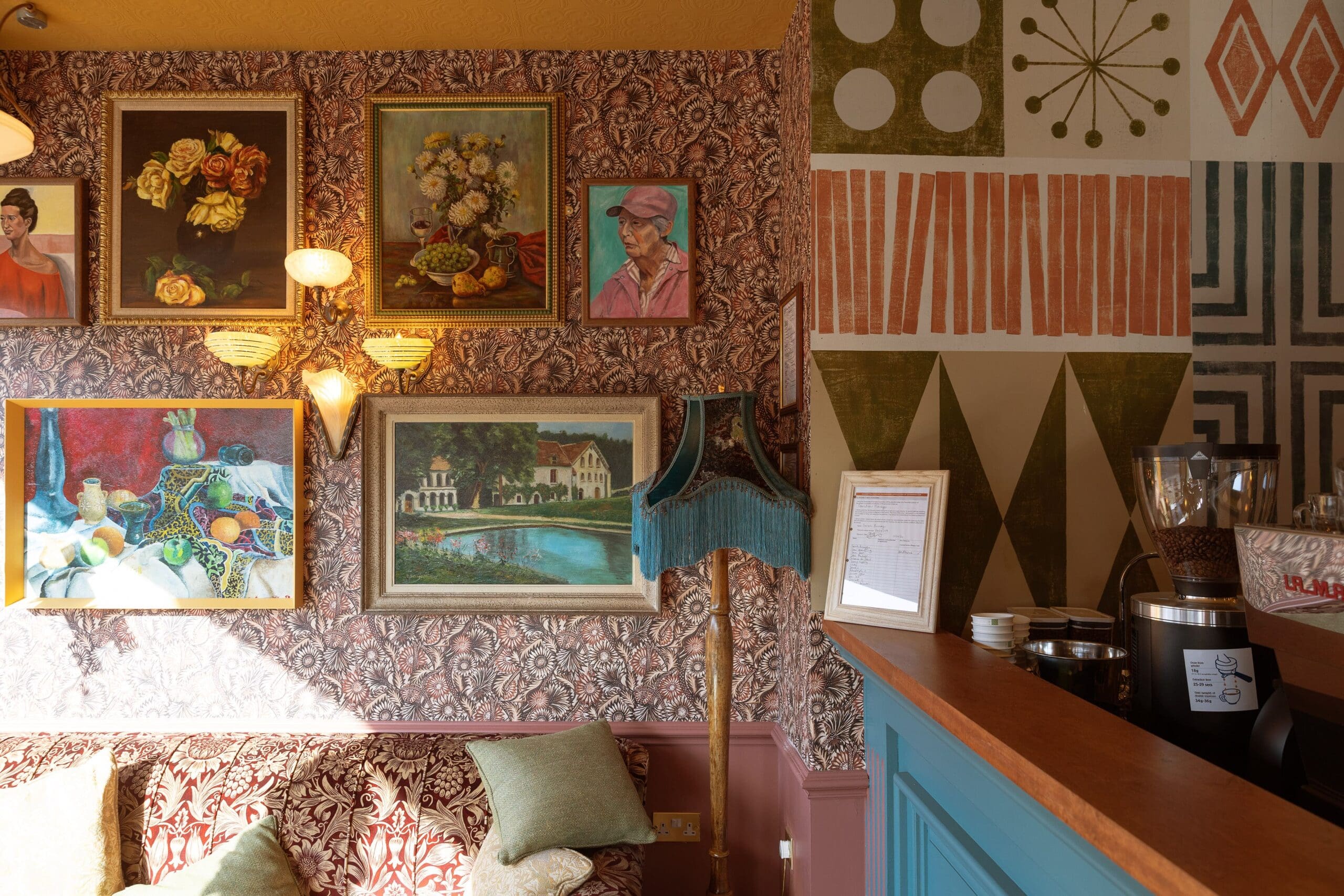 Eclectic café interior with patterned wallpaper, framed paintings, a vintage lamp, and a cushioned sofa. Sunlight streams across the Maggio turquoise counter with a coffee machine, enhancing the cozy and artistic atmosphere.