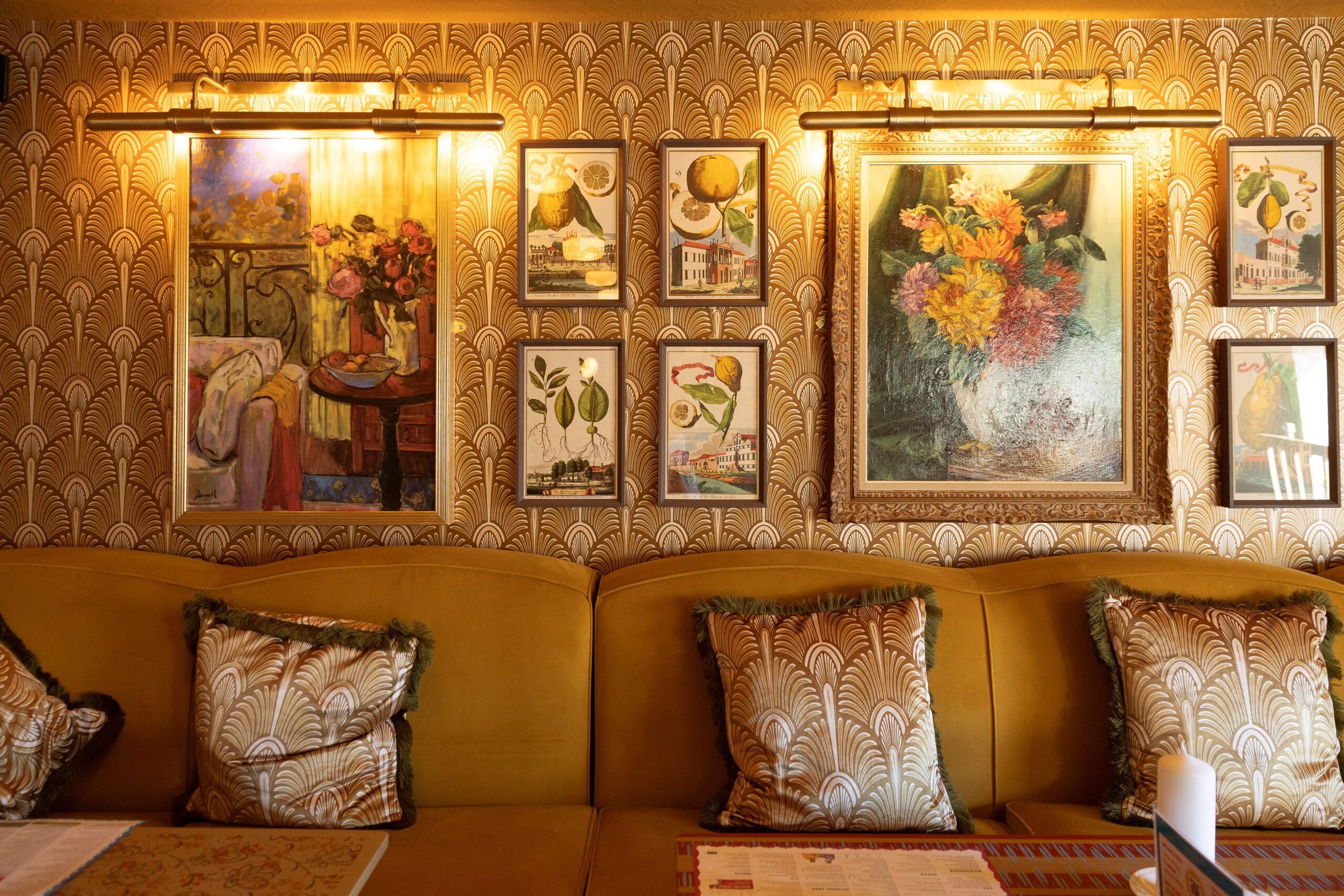 A cozy yellow Maggio couch with decorative pillows sits against a wall adorned with vintage floral paintings and botanical prints, all lit warmly by picture lights above.