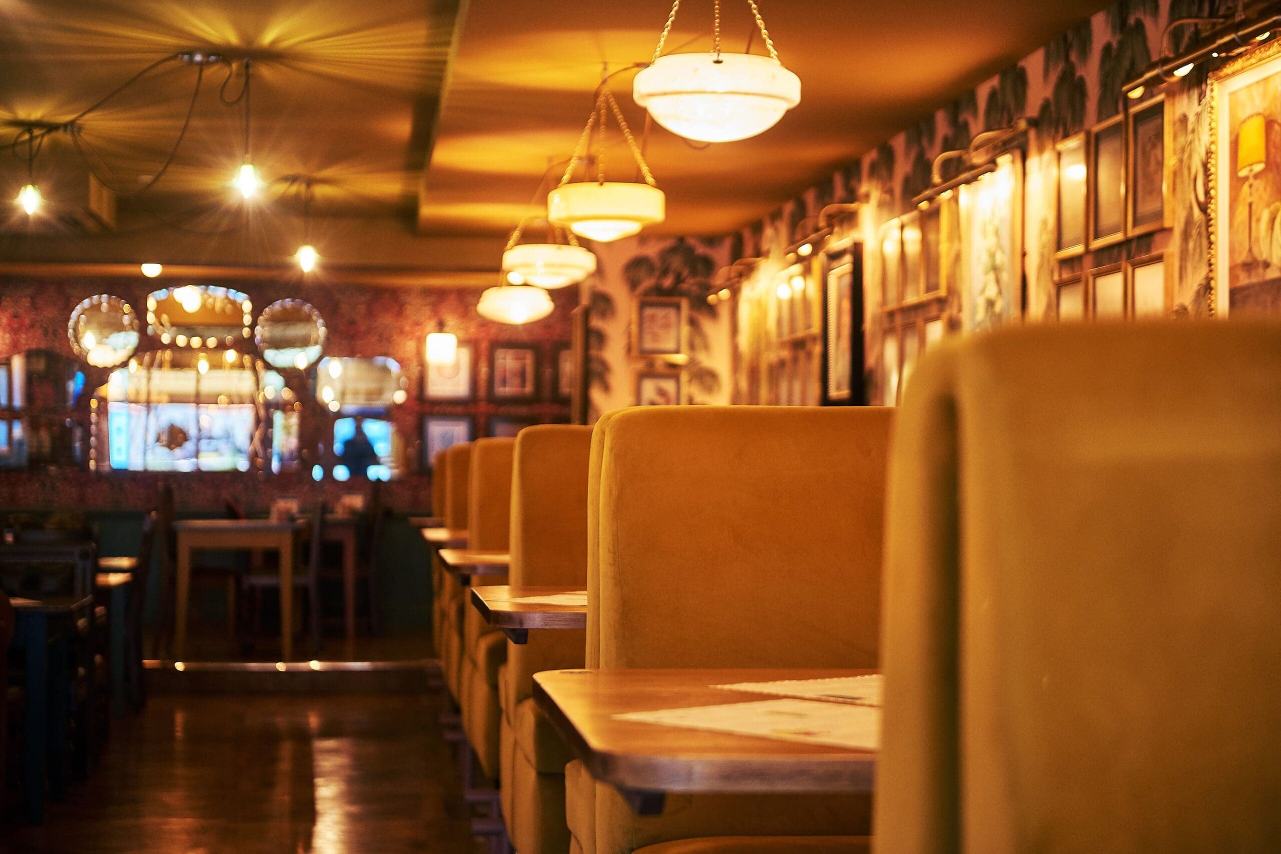 Warmly lit Hesco restaurant interior with empty yellow booths, wooden tables, decorative light fixtures, and framed pictures on patterned walls, creating a cozy and inviting atmosphere.