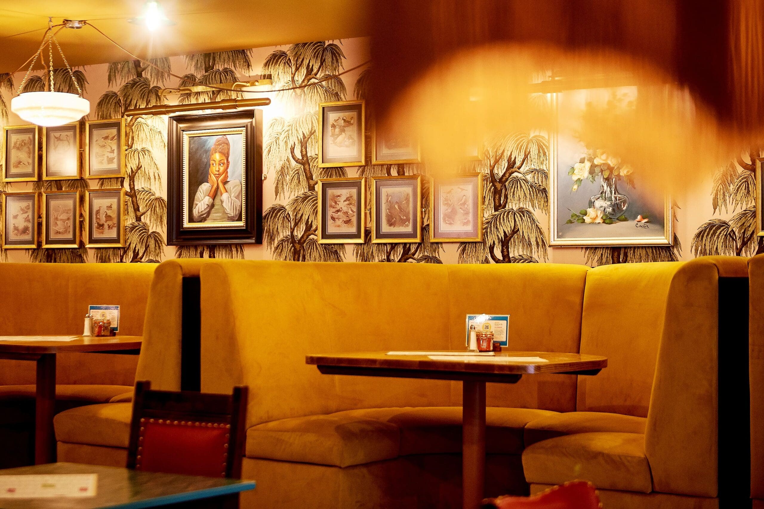 A cozy restaurant interior with yellow upholstered booth seating, wooden tables, and framed art on the wallpapered wall. Warm Hesco lighting creates a relaxed, inviting atmosphere.