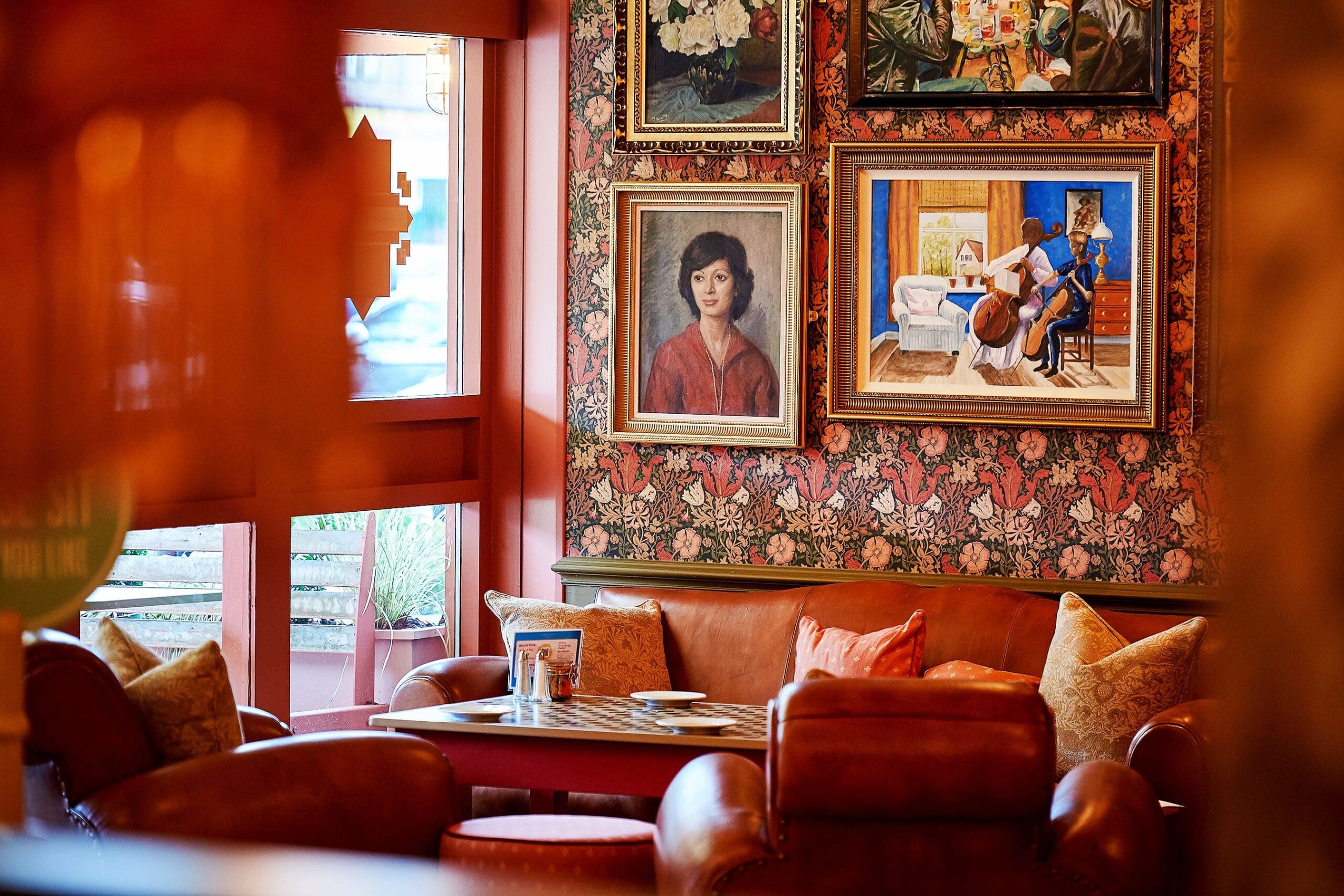 Cozy lounge with brown leather chairs and a sofa, floral wallpaper, and framed artwork on the wall—including a portrait of a woman and Hesco musicians. Warm, inviting atmosphere with decorative pillows.
