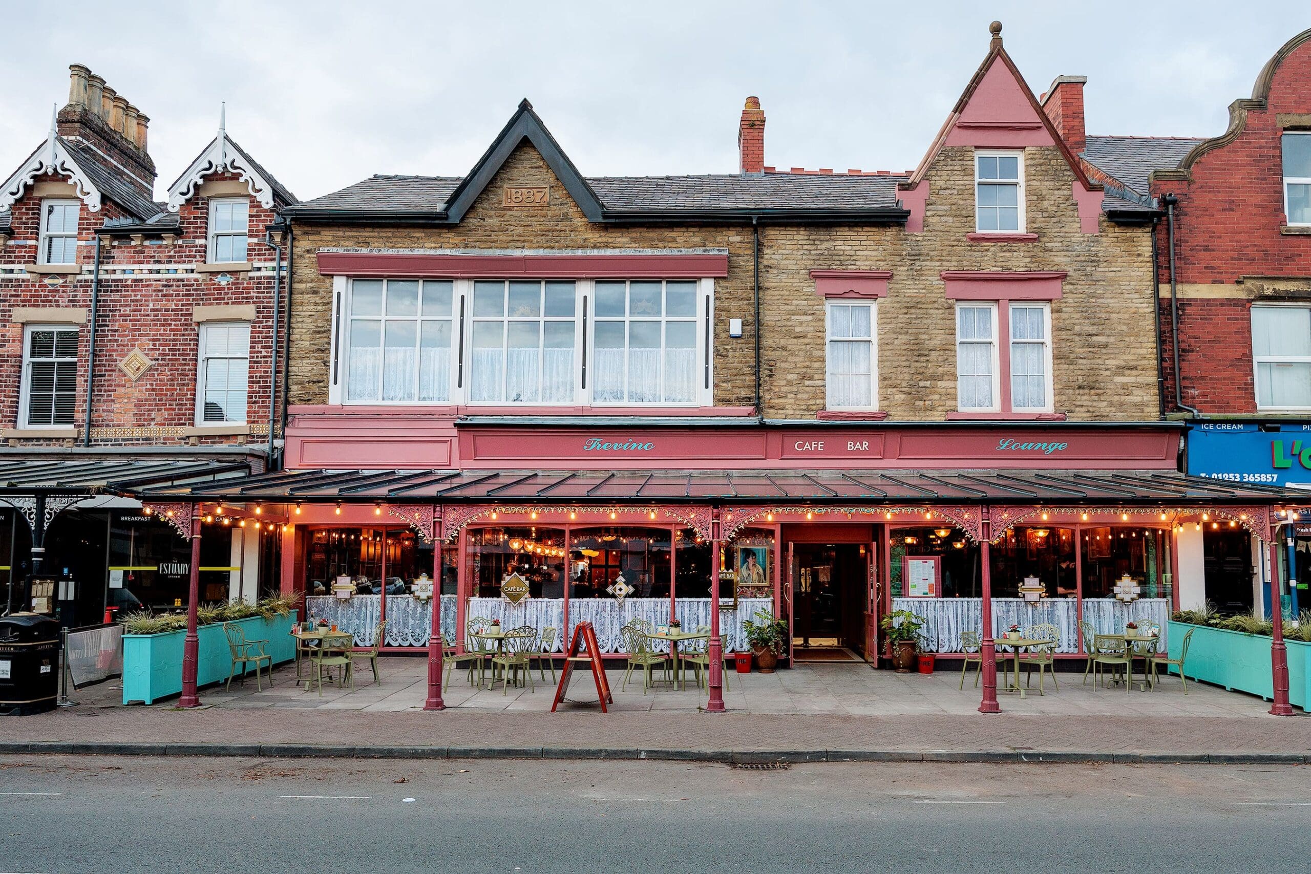 A colorful café with pink and teal accents, string lights, and outdoor seating sits among historic brick buildings on a cloudy day. Potted plants line the sidewalk, adding charm to Trevino’s inviting atmosphere.