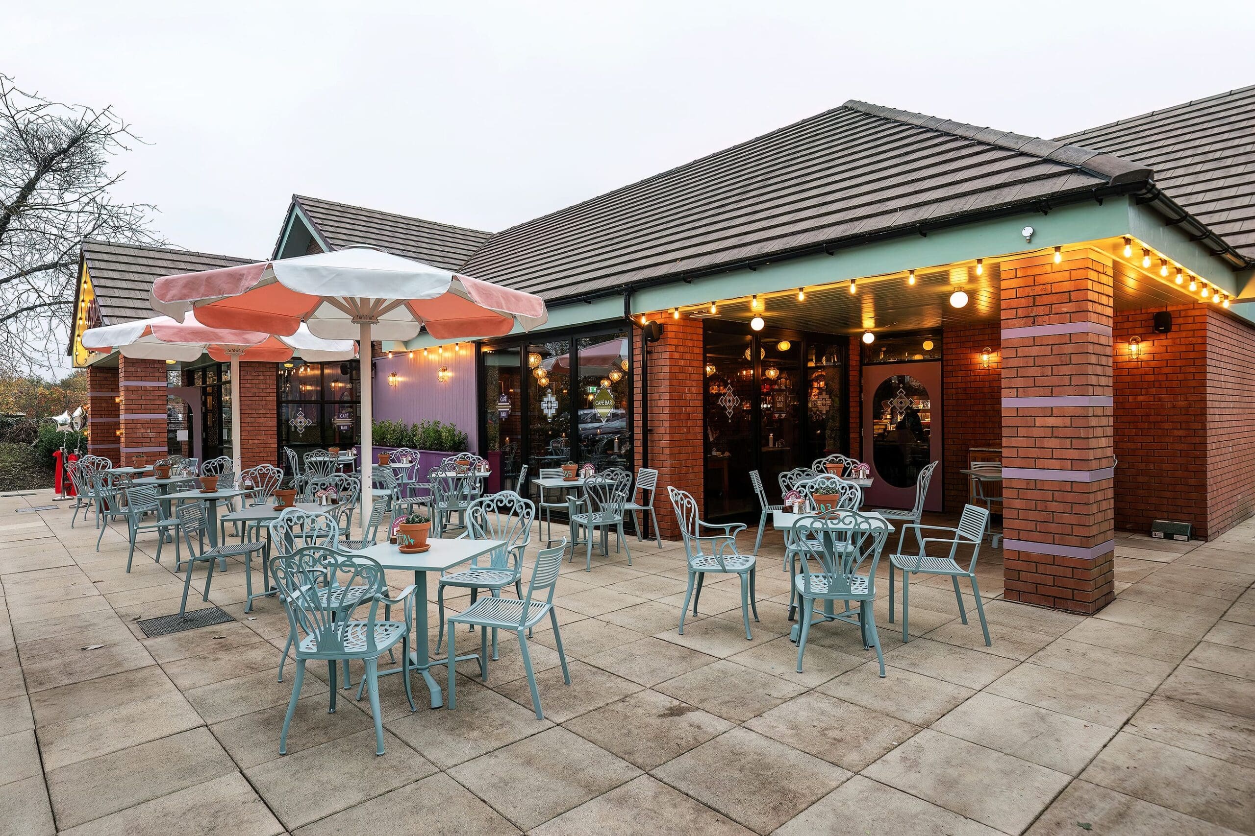 Outdoor seating area of a café with pastel blue metal tables and chairs, petalo-pink and white umbrellas, string lights under the roof, and large windows revealing a warmly lit interior. The setting appears calm and inviting.