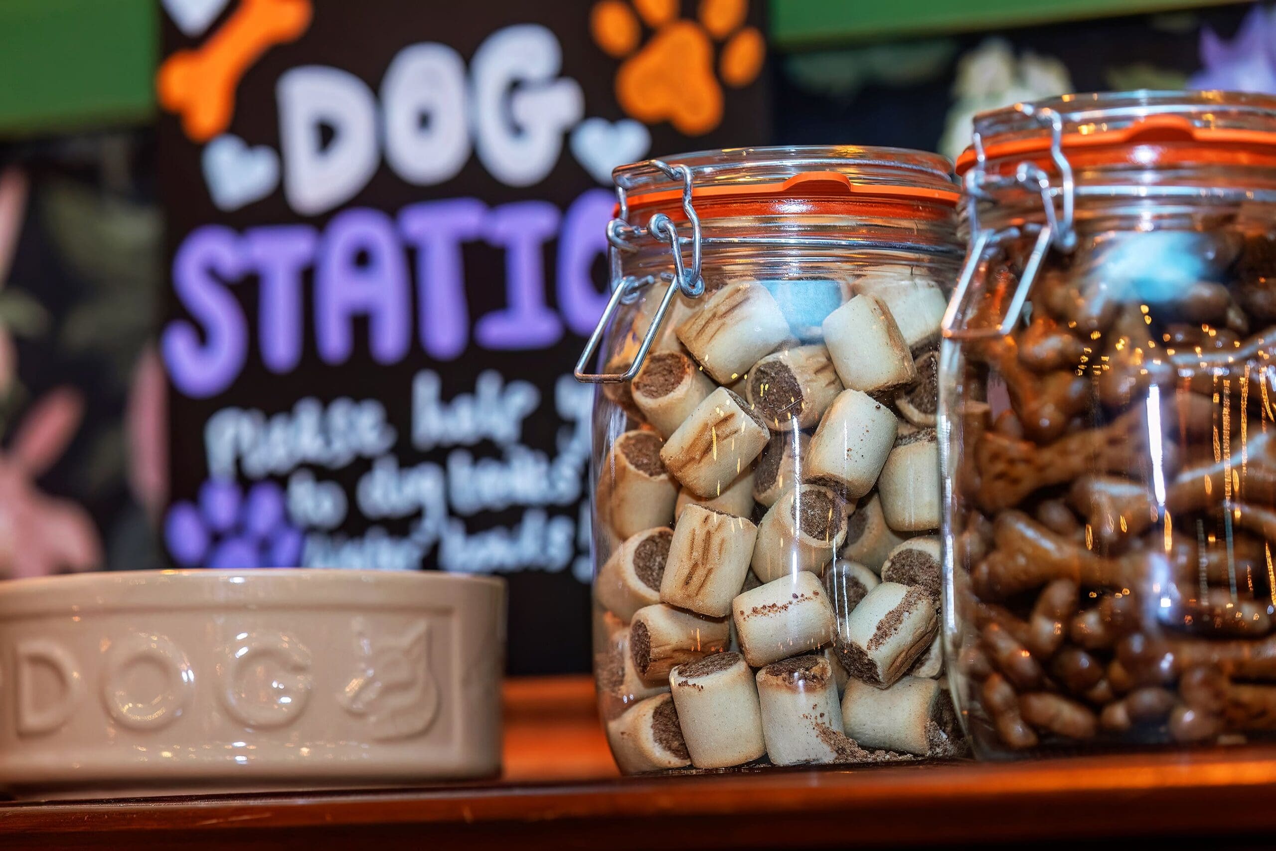 Two glass jars filled with Petalo dog treats sit next to a cream-colored dog bowl labeled "DOG," in front of a sign that reads "DOG STATION" with paw prints and playful text.