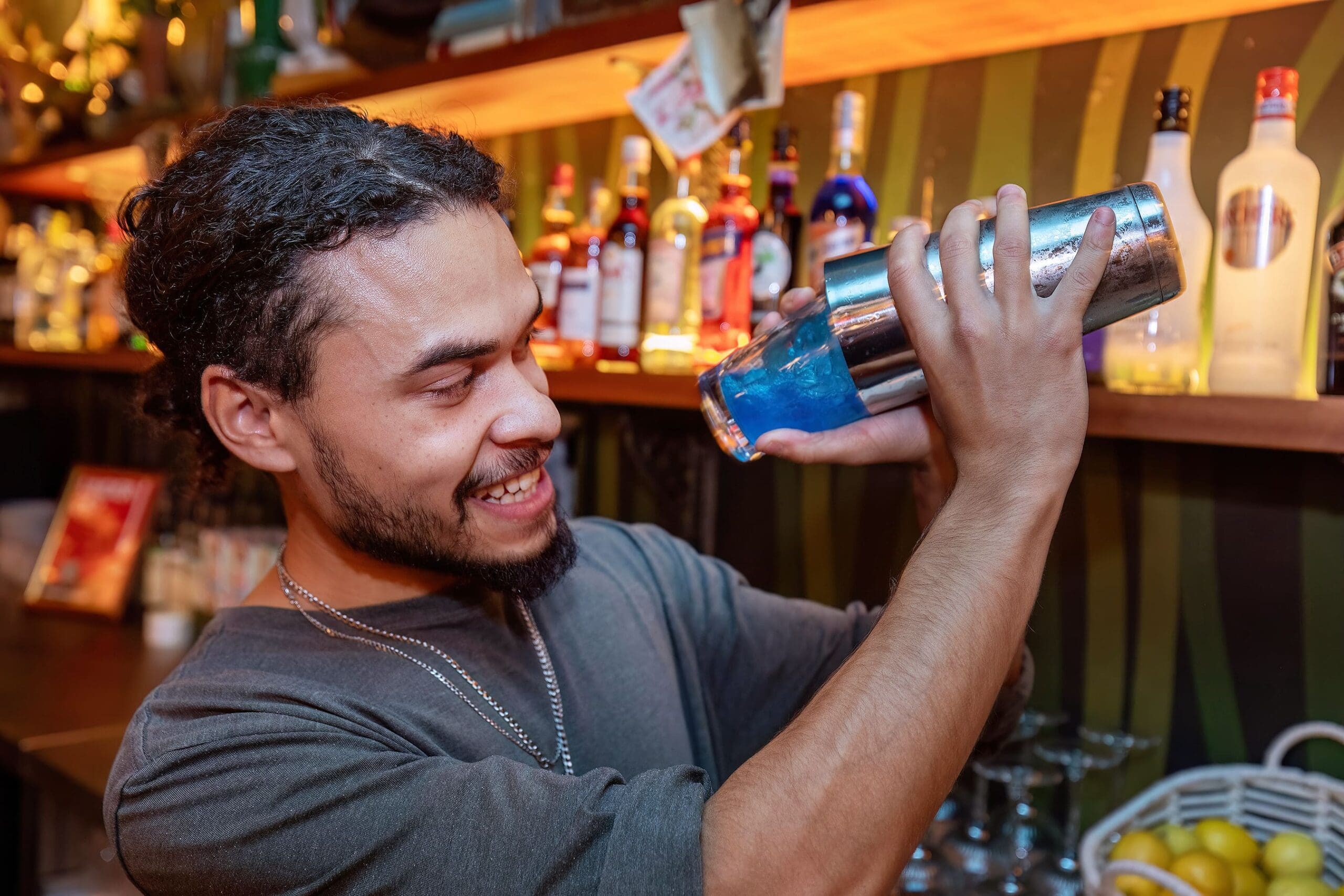 A bartender smiles while shaking a cocktail shaker with blue petalo liquid inside. Shelves of various bottles and barware are visible in the background.