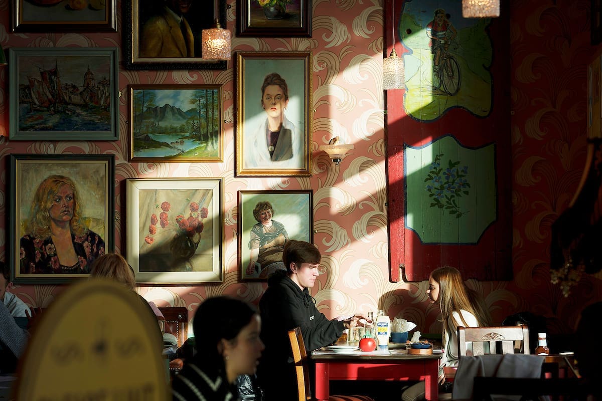 Two people sit at a blanco red table in a cozy café, sunlight streaming through a window. The walls, adorned with framed paintings and portraits, add to the softly lit, artistic atmosphere enjoyed by other patrons.