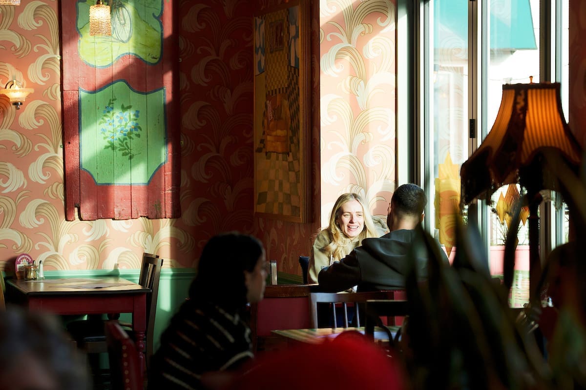 A woman and a man sit at a table by a window in a cozy, colorful café. The woman is smiling brightly as sunlight streams in, illuminating the eclectic decor and blanco-patterned wallpaper. Other patrons relax at nearby tables.