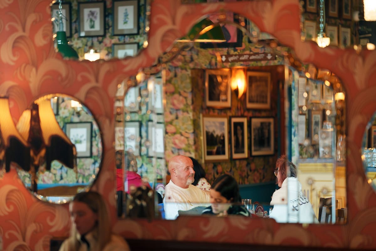 A decorative blanco mirror in a cozy, vintage-style restaurant reflects a smiling man and woman sitting at a table, engaged in conversation. Floral wallpaper, framed pictures, and warm lighting fill the background.