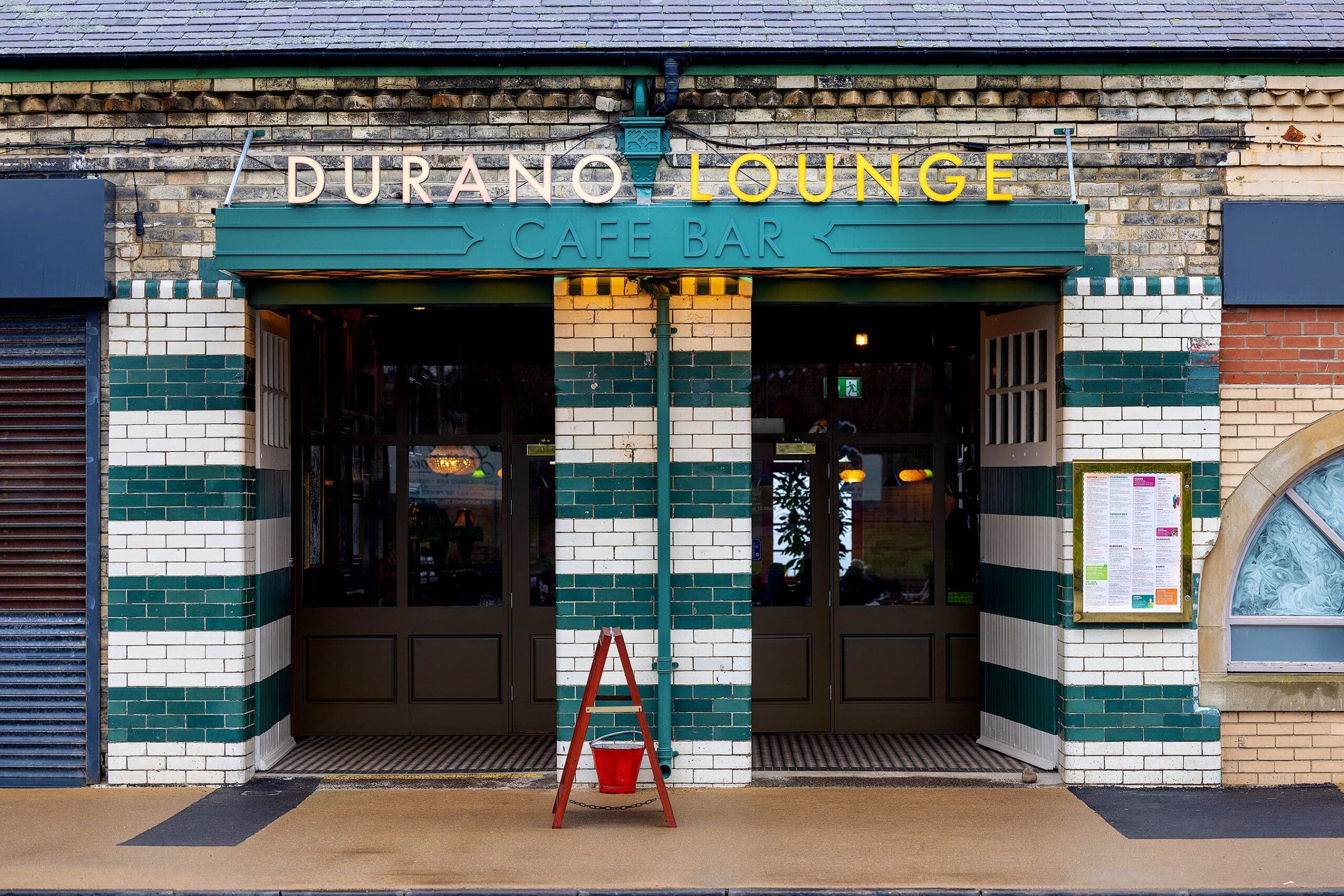 Front view of Durano Lounge café bar entrance with green and white tiled walls, double doors, and a yellow durano sign above. A red bucket sits on the pavement in front of the doorway.