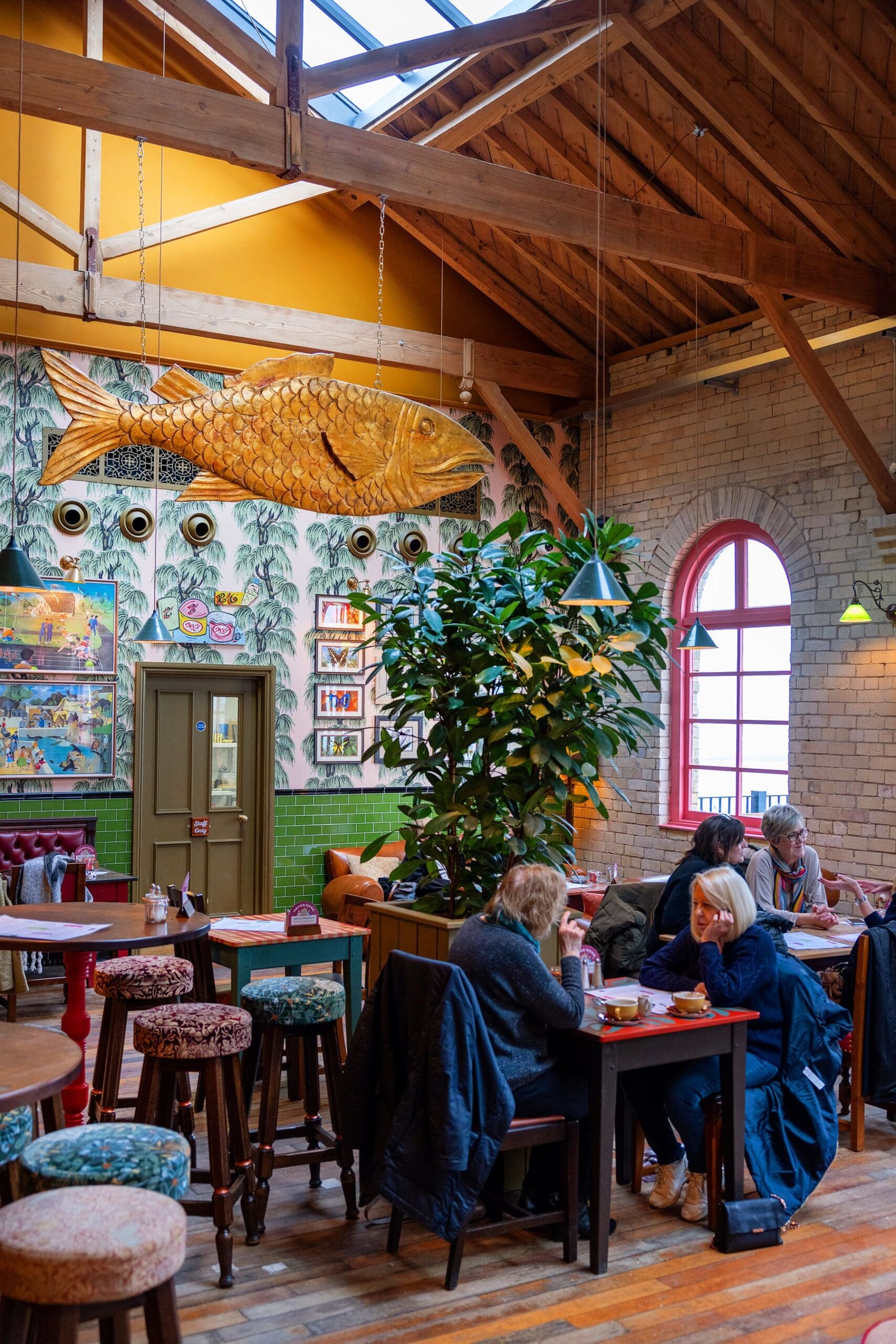 A cozy café with wooden beams, a large fish sculpture on the wall, green plants, and people sipping drinks under warm lighting—the perfect spot to try the signature durano pastry.