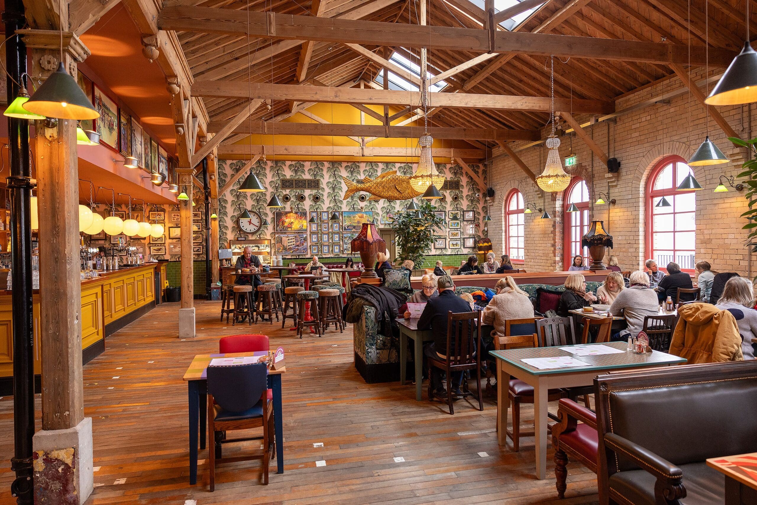 A spacious, lively restaurant with wooden beams, eclectic decor, large windows, and groups of people dining at tables. The bar area is on the left, and the warm durano atmosphere is both inviting and vibrant.