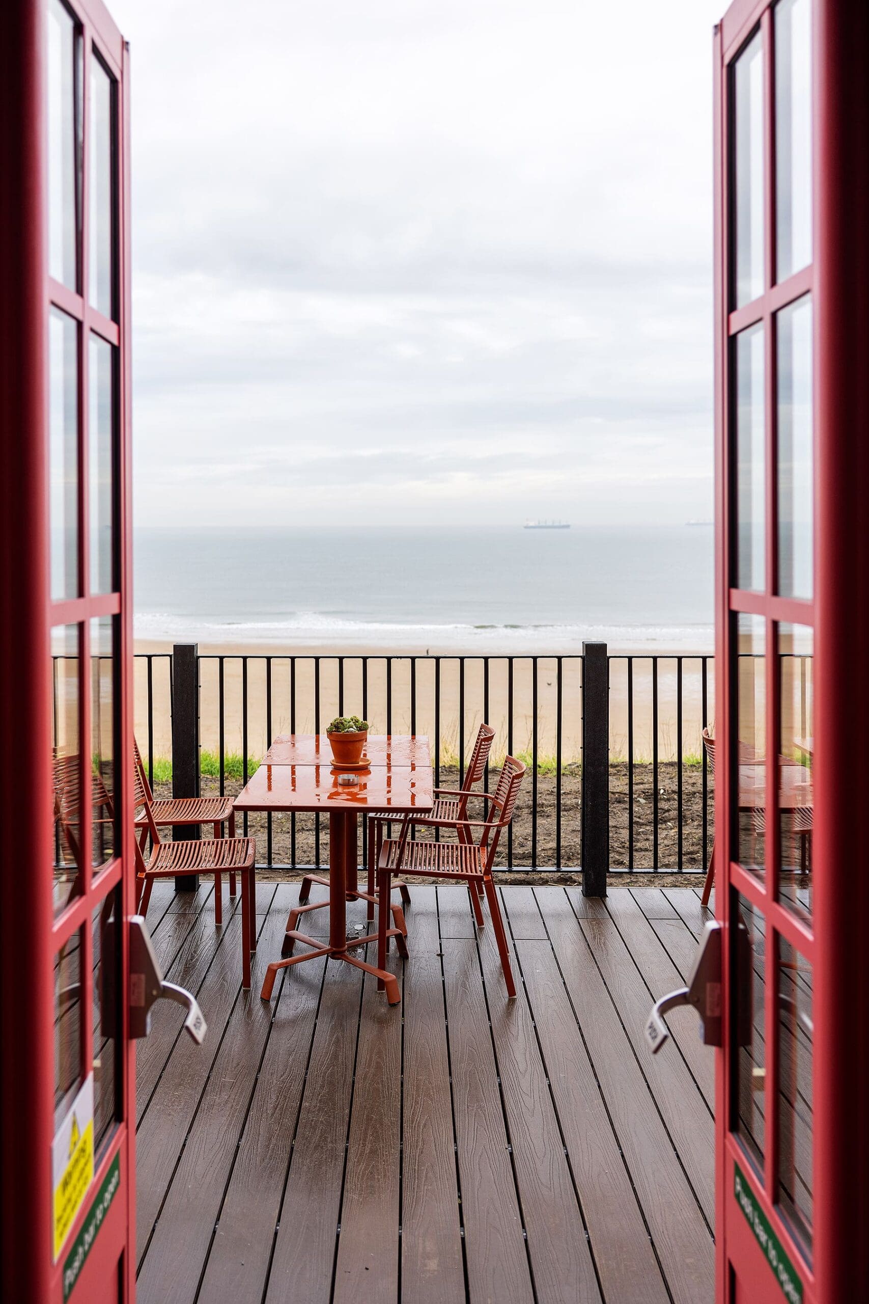 A view through open red doors to a patio with a table, four chairs, and a potted plant—featuring a touch of durano—overlooking a sandy beach and calm sea under a cloudy sky.