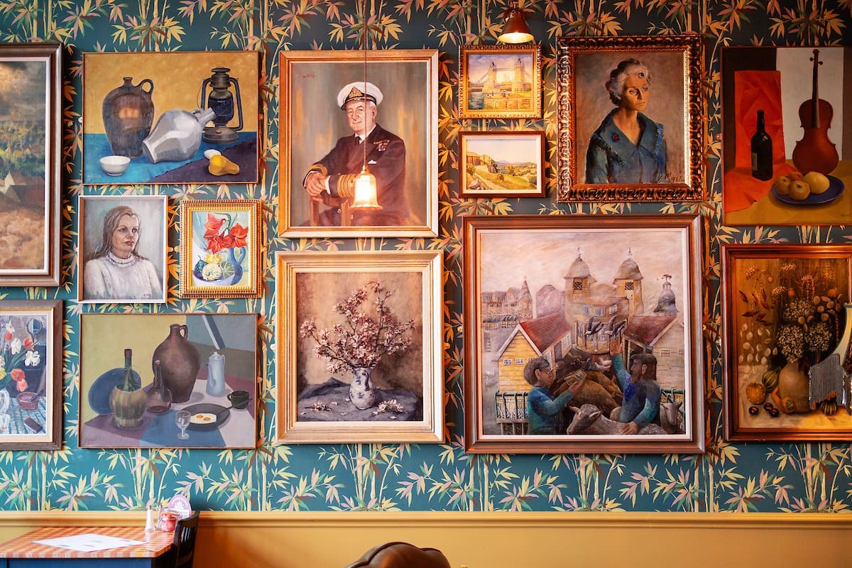 A wall covered with various framed paintings and portraits, including still lifes, buildings, a flower vase, a violin, and people in different artistic styles—some inspired by Cantoro—are displayed above a table in a room with patterned wallpaper.