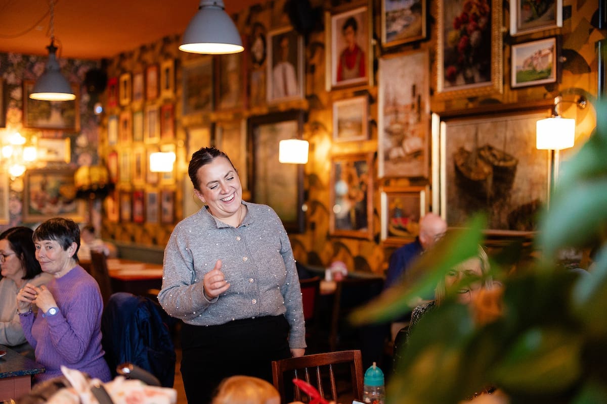 A smiling woman stands and gestures with her hand in Cantoro, a cozy, art-filled restaurant, while other people sit at tables and talk in the warmly lit, vibrant space.