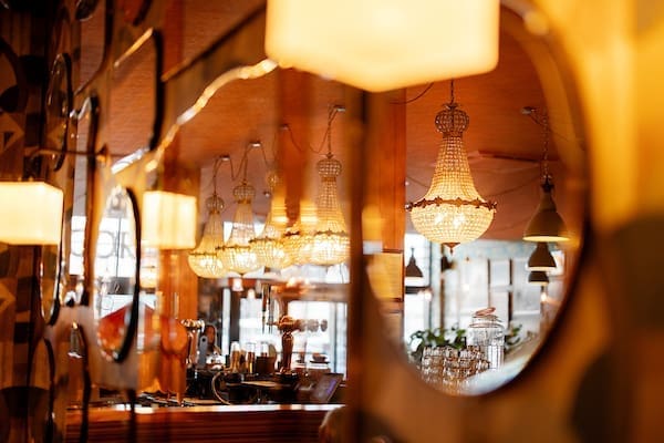 Warmly lit Cantoro restaurant interior reflected in decorative mirrors, featuring ornate chandeliers, hanging lamps, bar taps, and rows of glasses, creating a cozy and inviting atmosphere.