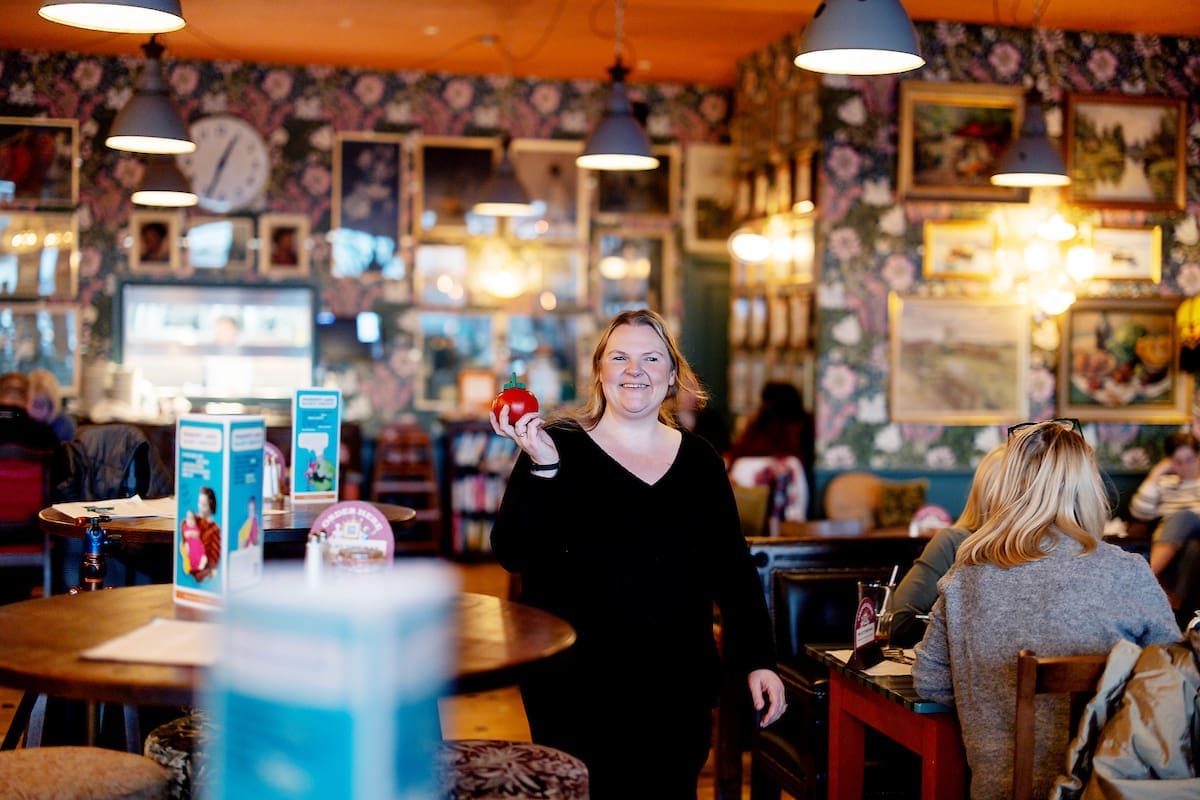 A woman in a black top smiles and holds a red apple while standing in Cantoro, a cozy, brightly lit café with floral wallpaper and framed pictures. Other people sit at tables in the background.