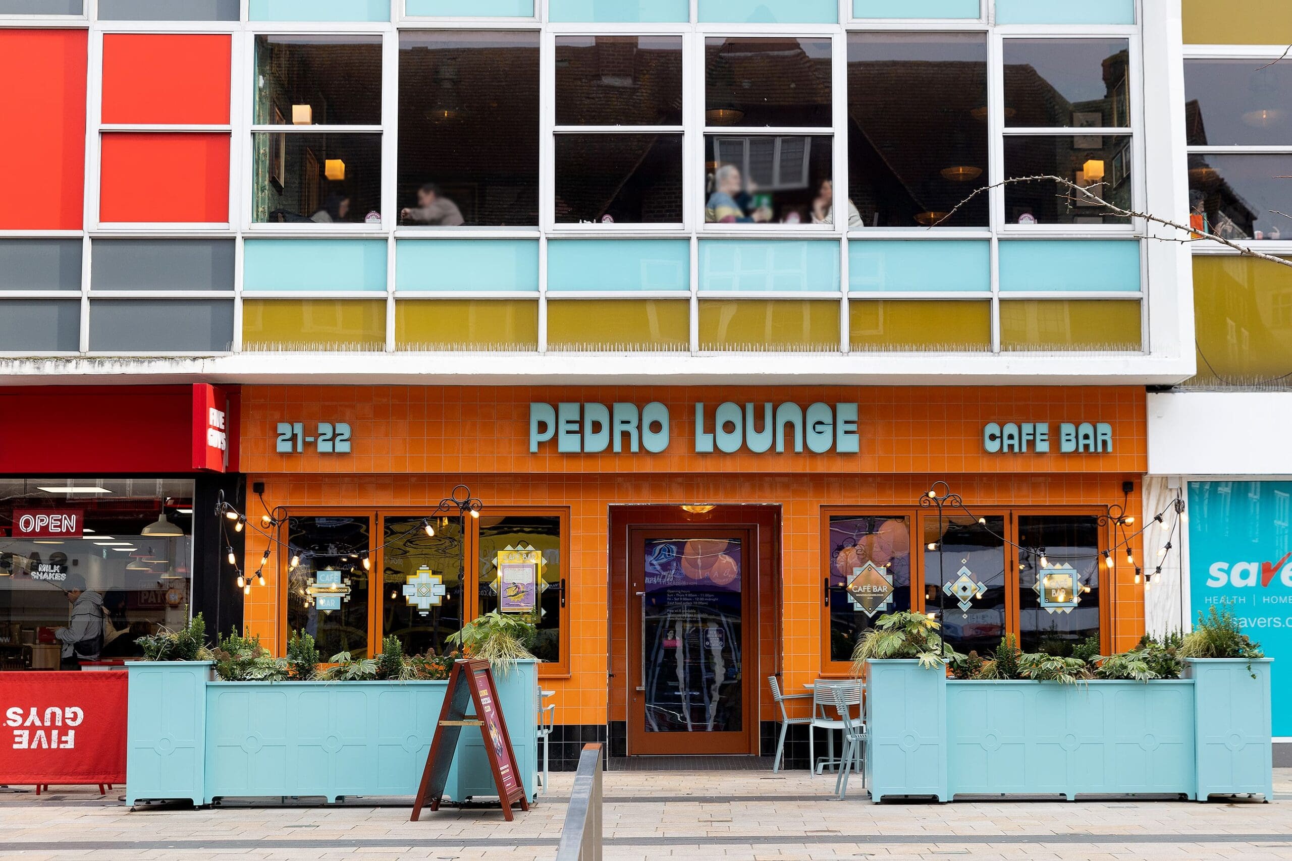 The exterior of Pedro Lounge café bar features bright orange tiles, large windows above, teal planters with greenery, and outdoor tables on a paved walkway. "Pedro Lounge" and "Cafe Bar" are written above the entrance, highlighting its inviting pedro style.