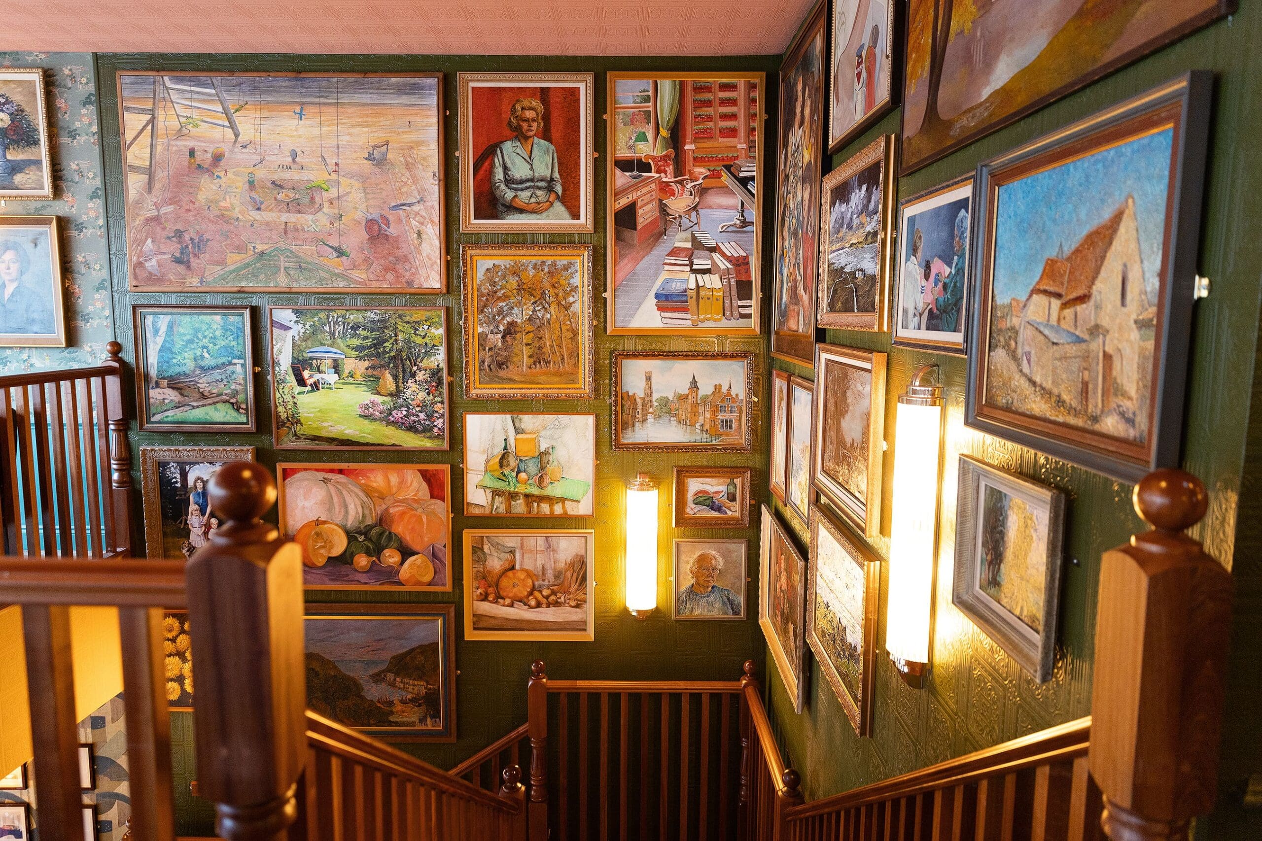 A staircase with wooden railings is surrounded by numerous framed paintings on green walls, featuring portraits, landscapes, and still lifes—some inspired by Pedro. Two vertical wall lights illuminate the colorful art display.