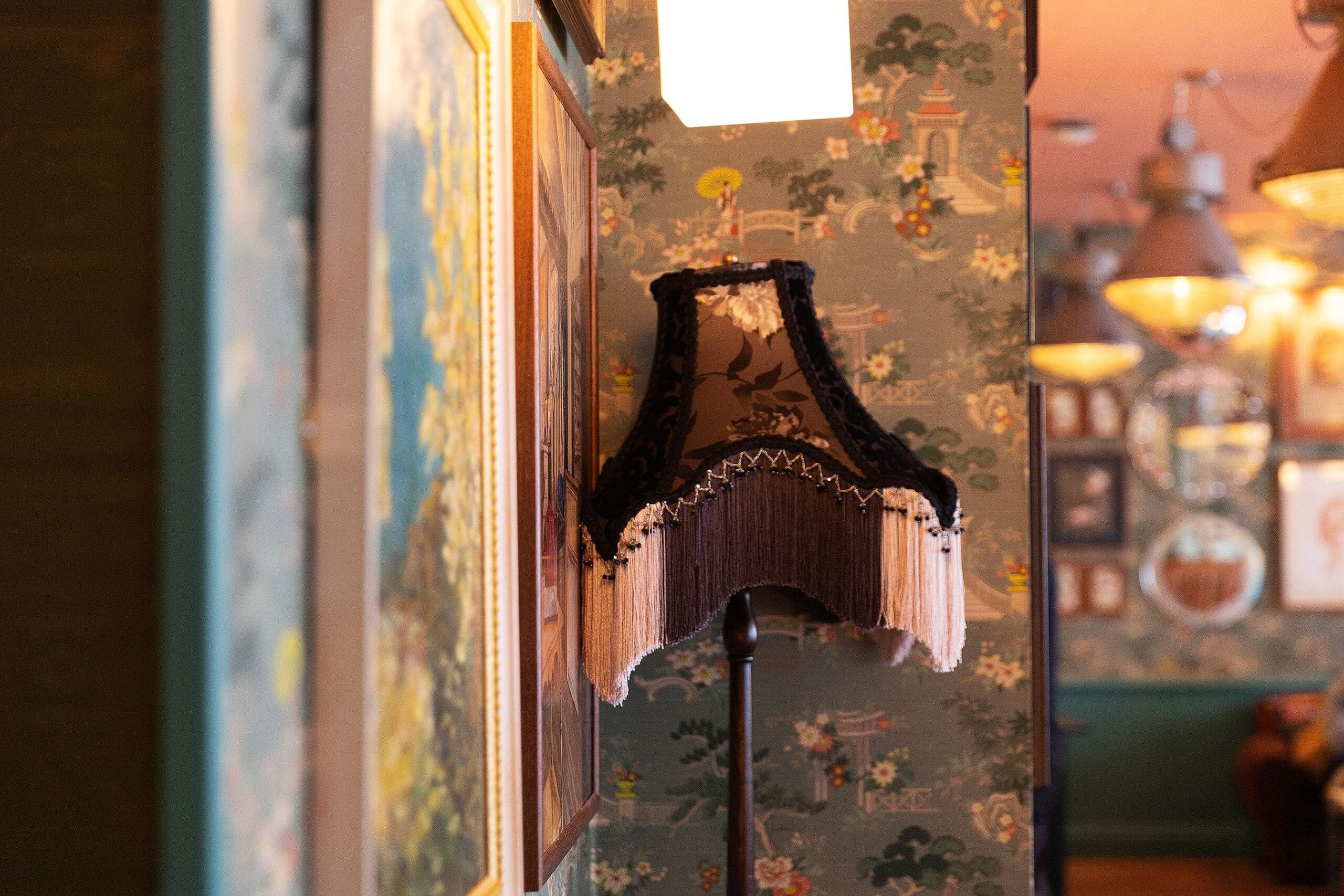 A vintage black lampshade with fringe stands against a decorative wallpapered wall, surrounded by framed art and mirrors in Pedro’s cozy, warmly lit room.