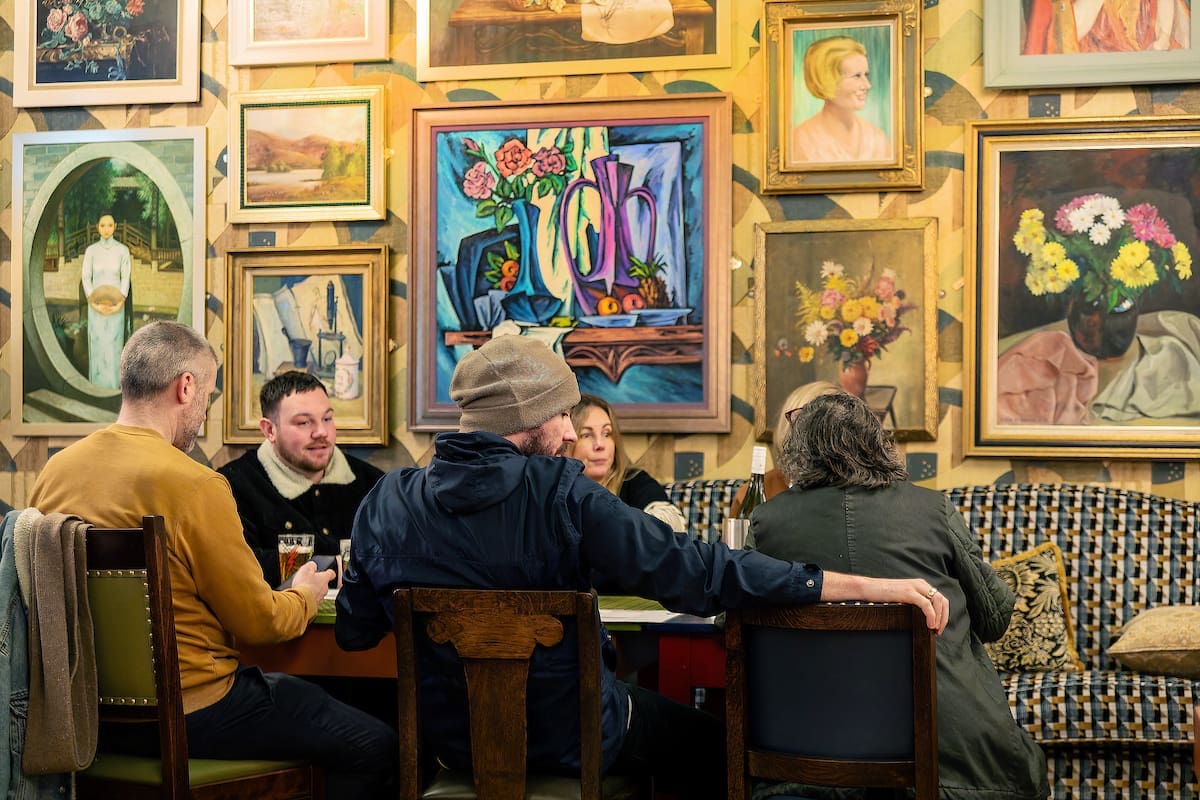 A group of five people sit around a table talking and drinking in a cozy room filled with colorful framed paintings—mostly portraits, still lifes, and even a calvero—hanging on the patterned wall behind them.